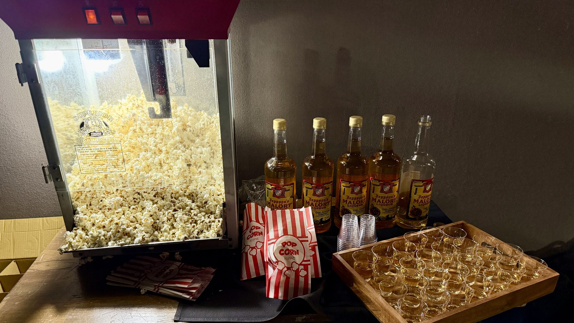 Popcorn machine filled with popcorn, red-and-white popcorn bags, five bottles of Jeppson's Malört liqueur, and a wooden tray with many small filled plastic cups on a table.