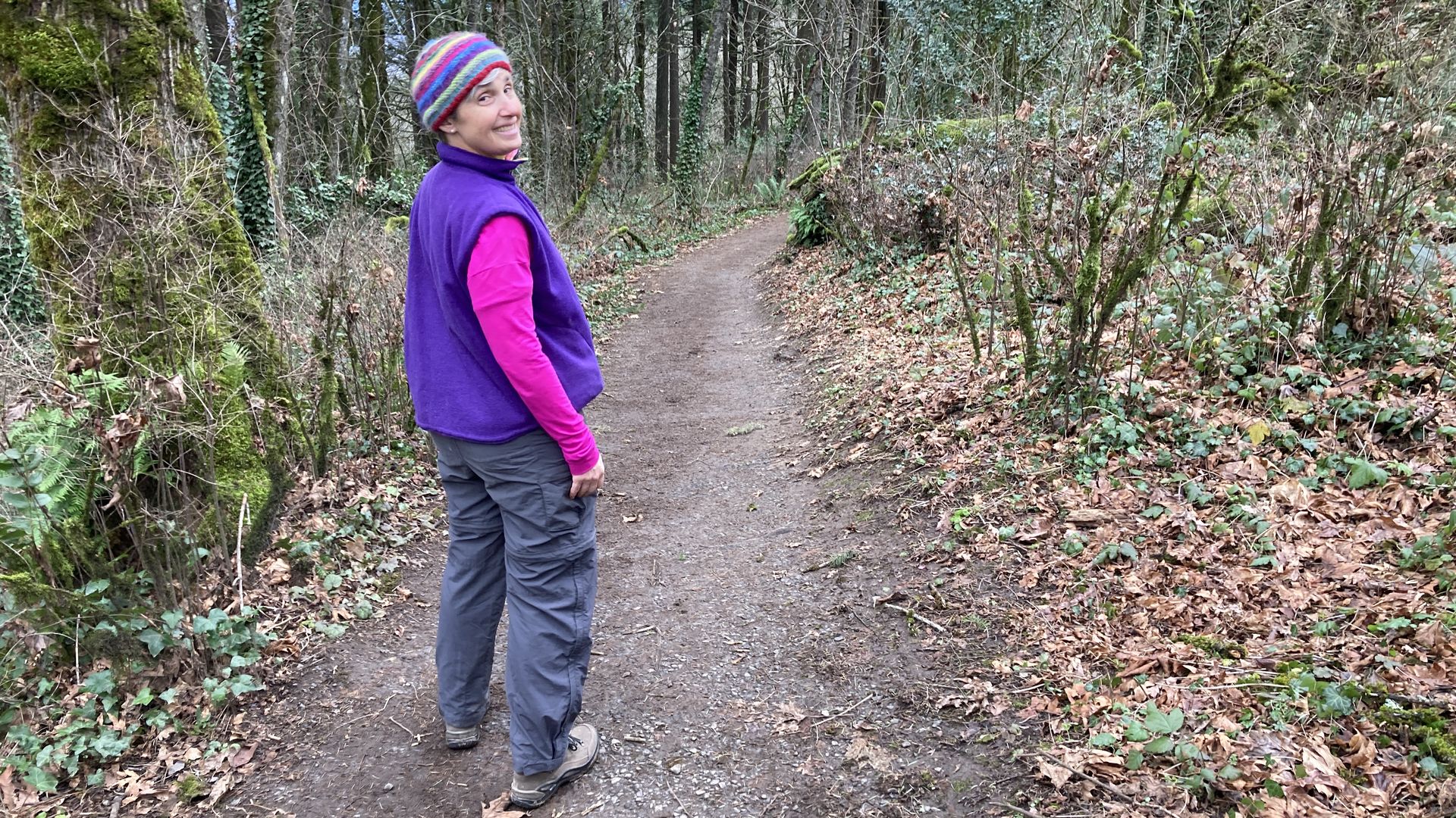 A woman in a bright pink shirt, purple vest and striped hat looks back at the camera as she stands on a dirt trail through the woods.