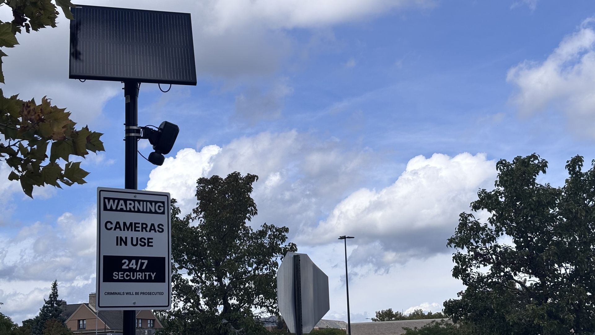 Tall street light with a large solar panel on a pole. A warning sign reads WARNING CAMERAS IN USE 24/7 SECURITY. Blue sky with scattered clouds and trees in the background.