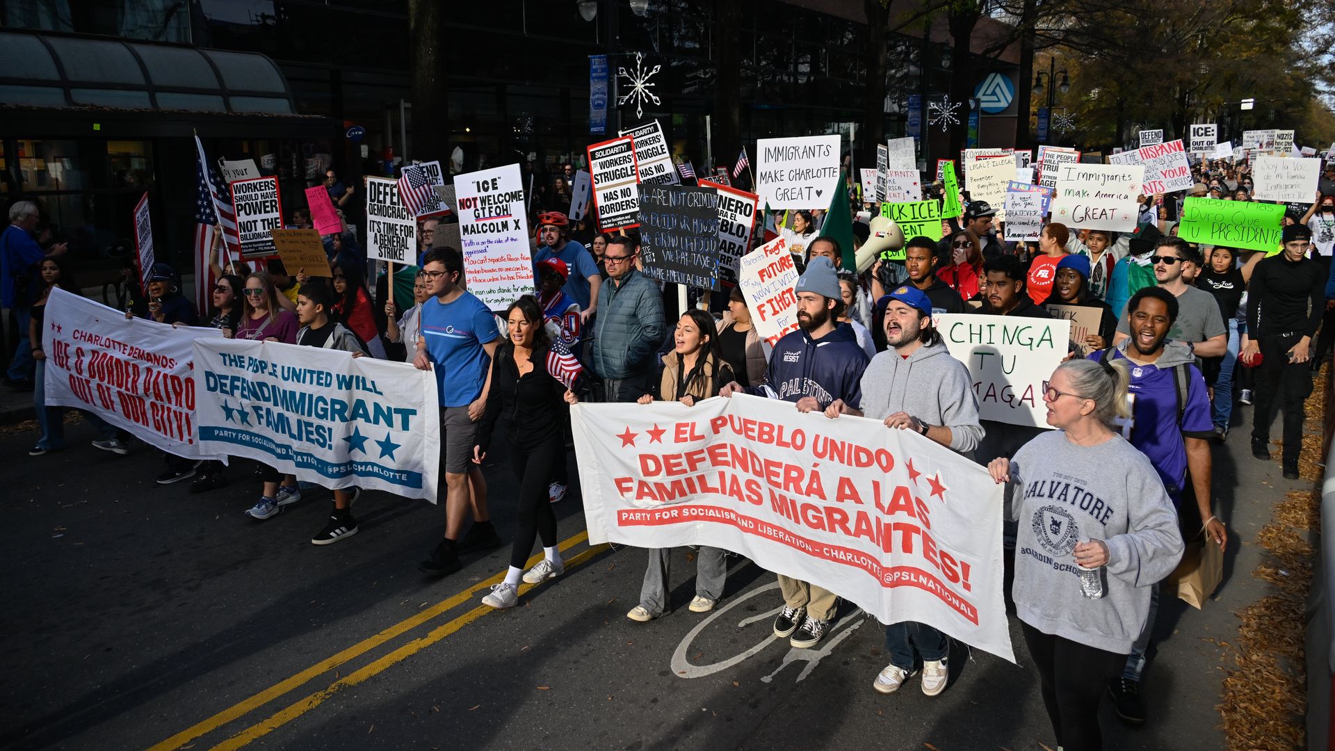 Diverse group of protesters marching in street holding banners and signs advocating for immigrant rights, including Spanish and English messages supporting immigrant families and opposing ICE.
