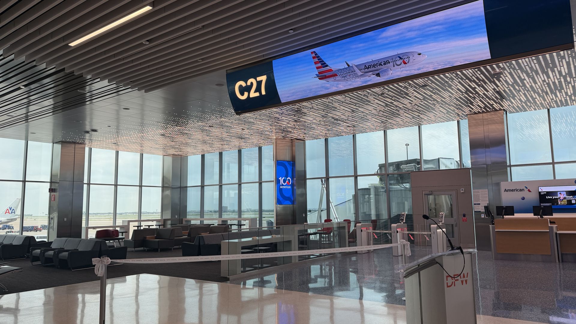 Inside a modern airport terminal at gate C27, a large sign shows C27 beside a digital banner of an American Airlines plane. Glass walls, gray seating, and a bright concourse with daylight.