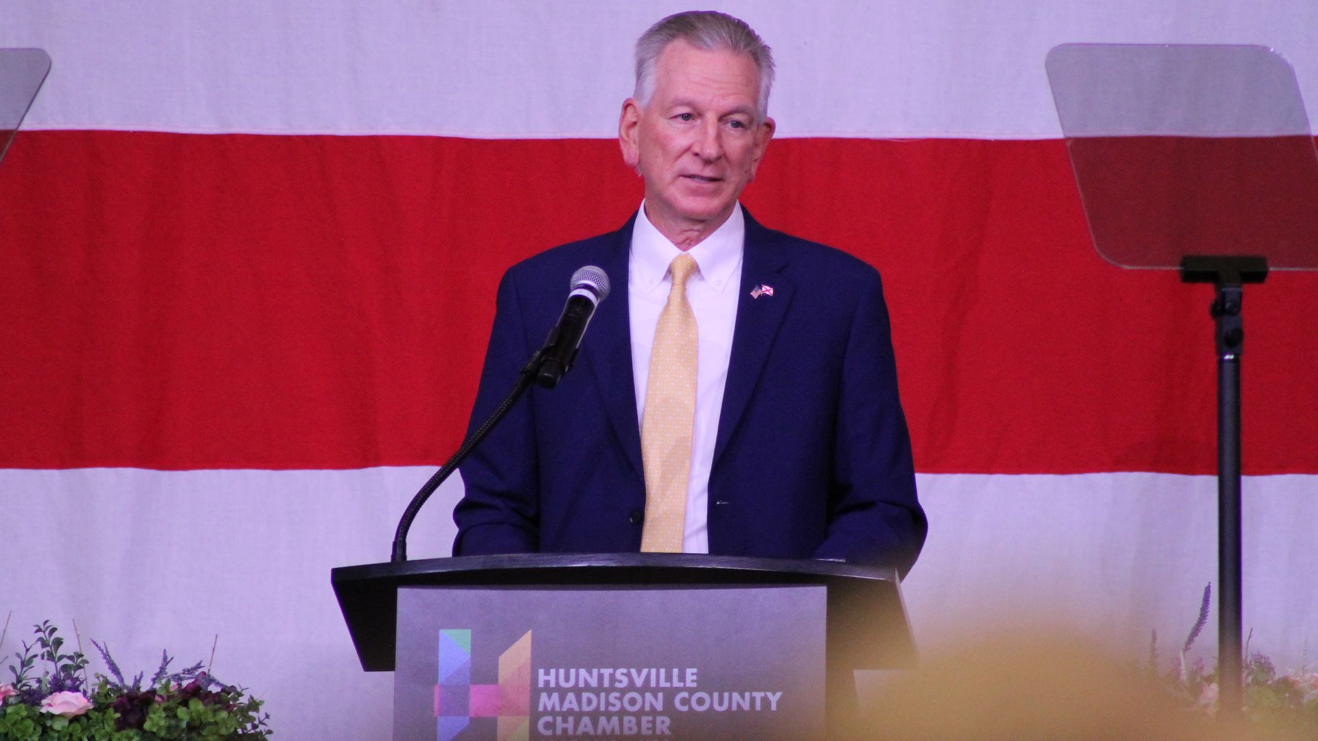 A man in a navy suit and yellow tie speaks at a podium with the Huntsville Madison County Chamber logo, in front of a large red and white striped backdrop.