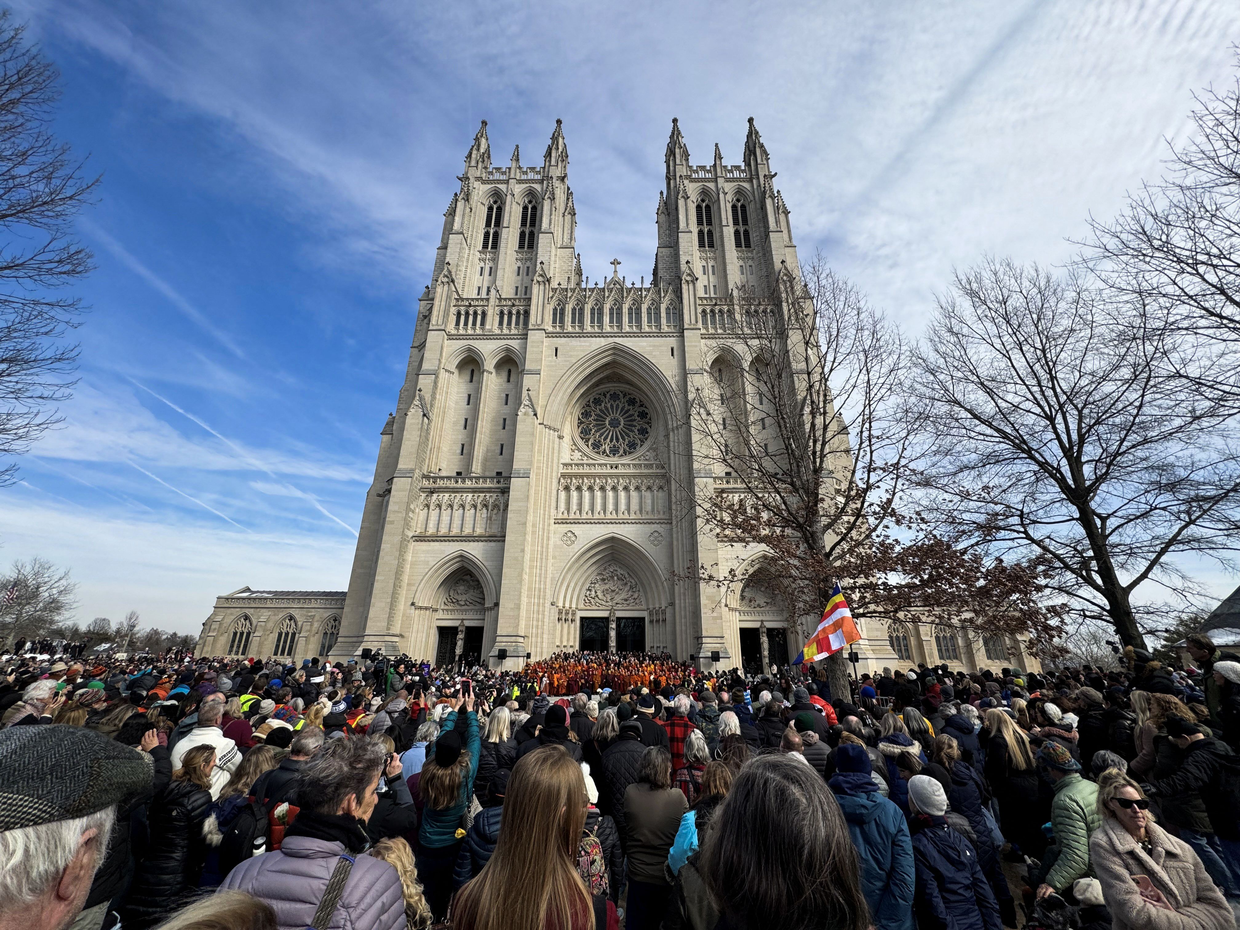 Large crowd gathered in front of a grand Gothic cathedral with twin towers under a partly cloudy blue sky during an outdoor event.