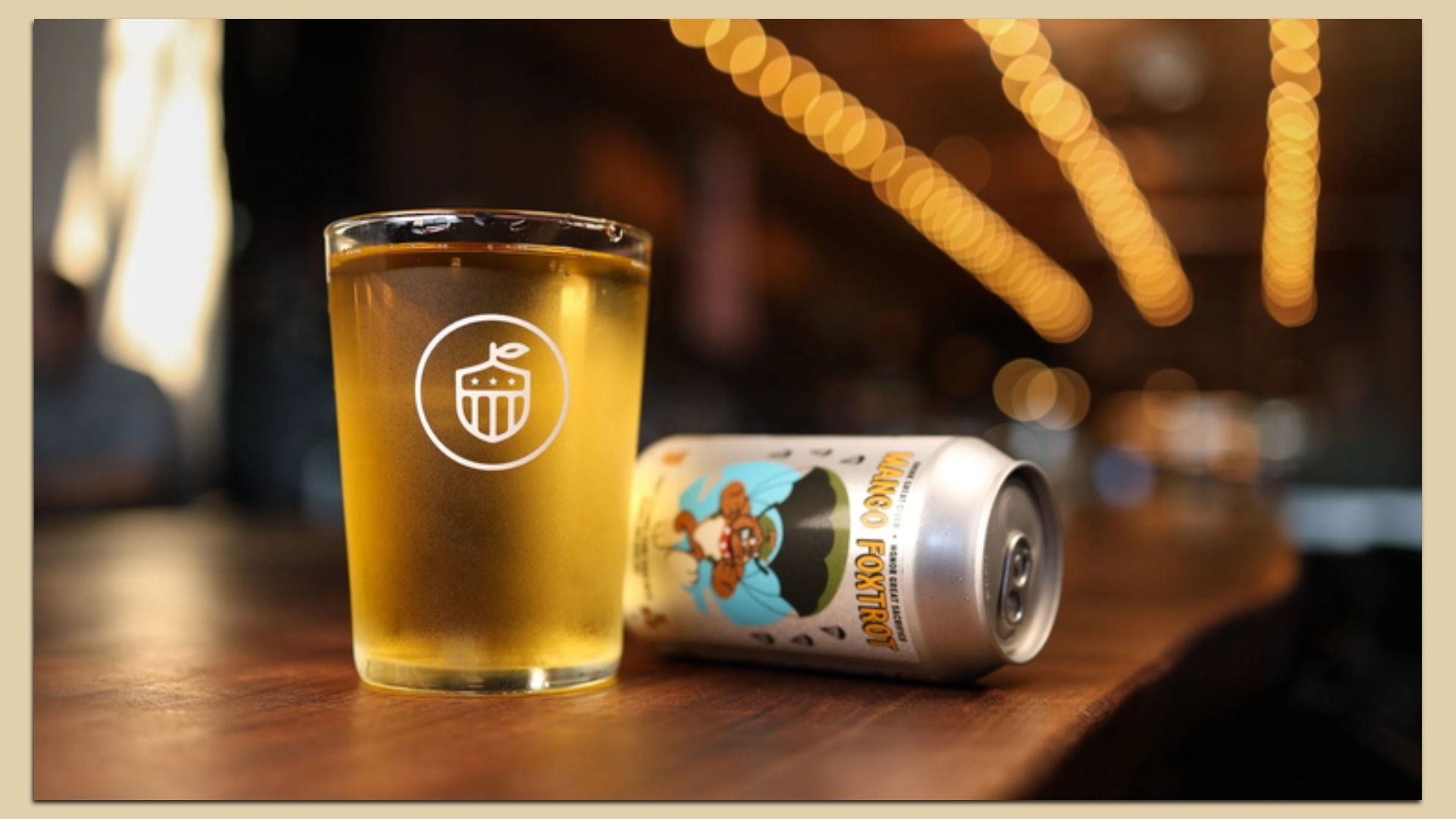 Golden beer in a tall glass on a wooden bar, beside a colorful can. The glass shows a white circular cupcake logo; warm string lights blur into a soft bokeh background.
