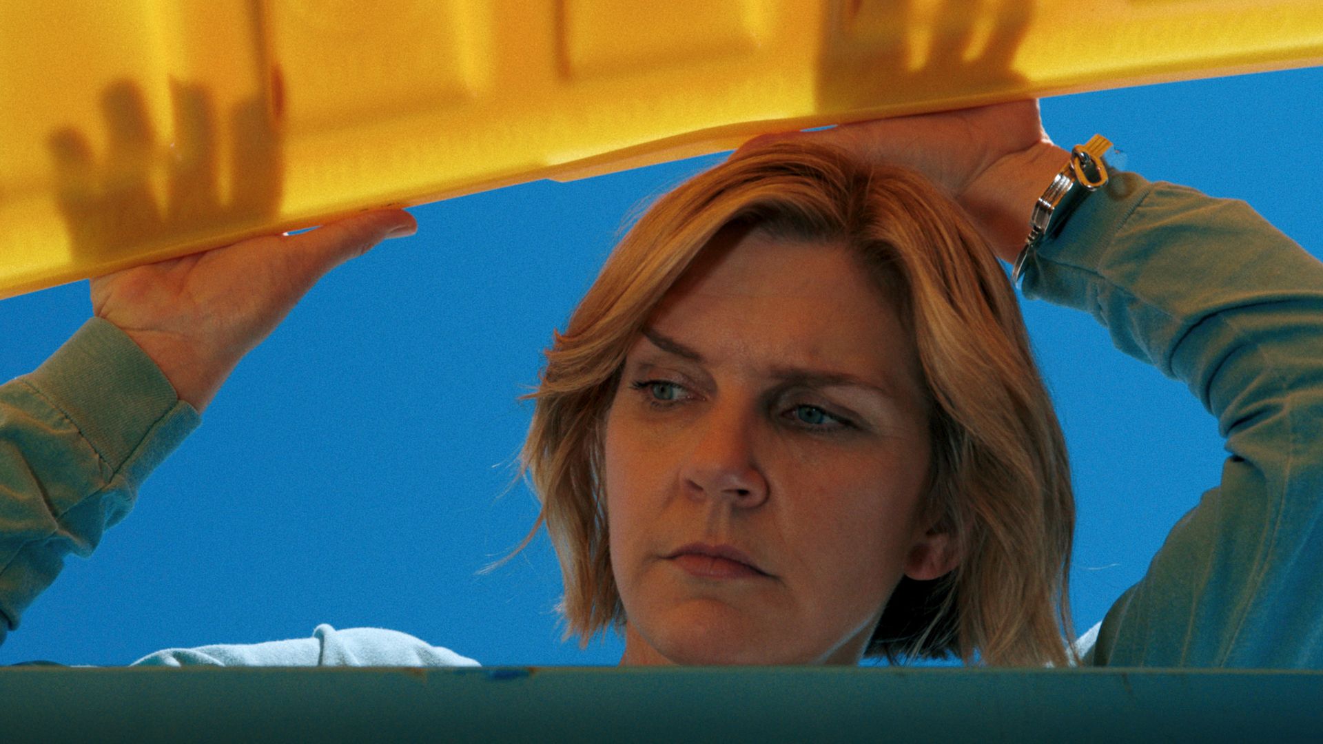 A woman lifts a yellow lid of a dumpster. The background is bright beautiful blue.