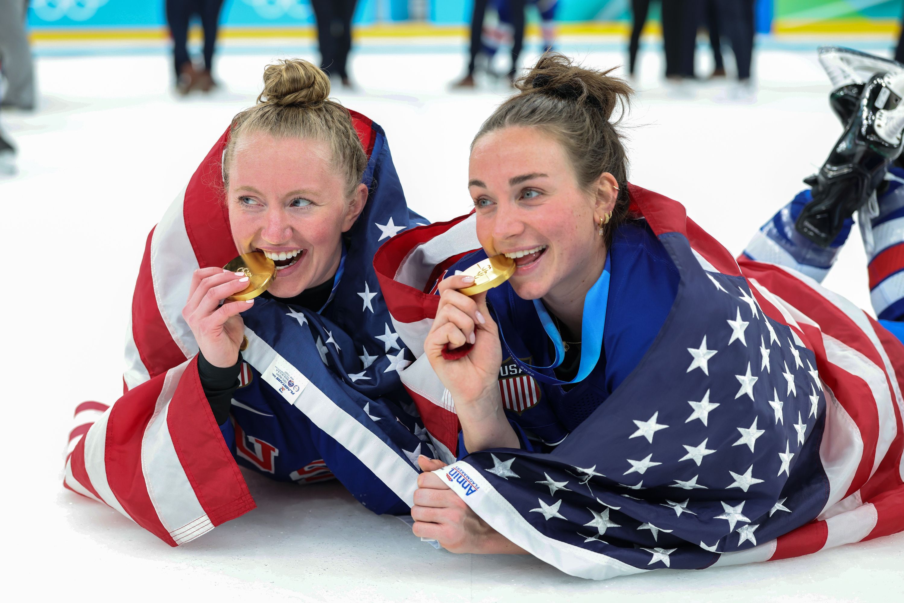 Two female athletes wrapped in American flags lie on ice biting gold medals, celebrating a victory in blue USA uniforms and ice skates.