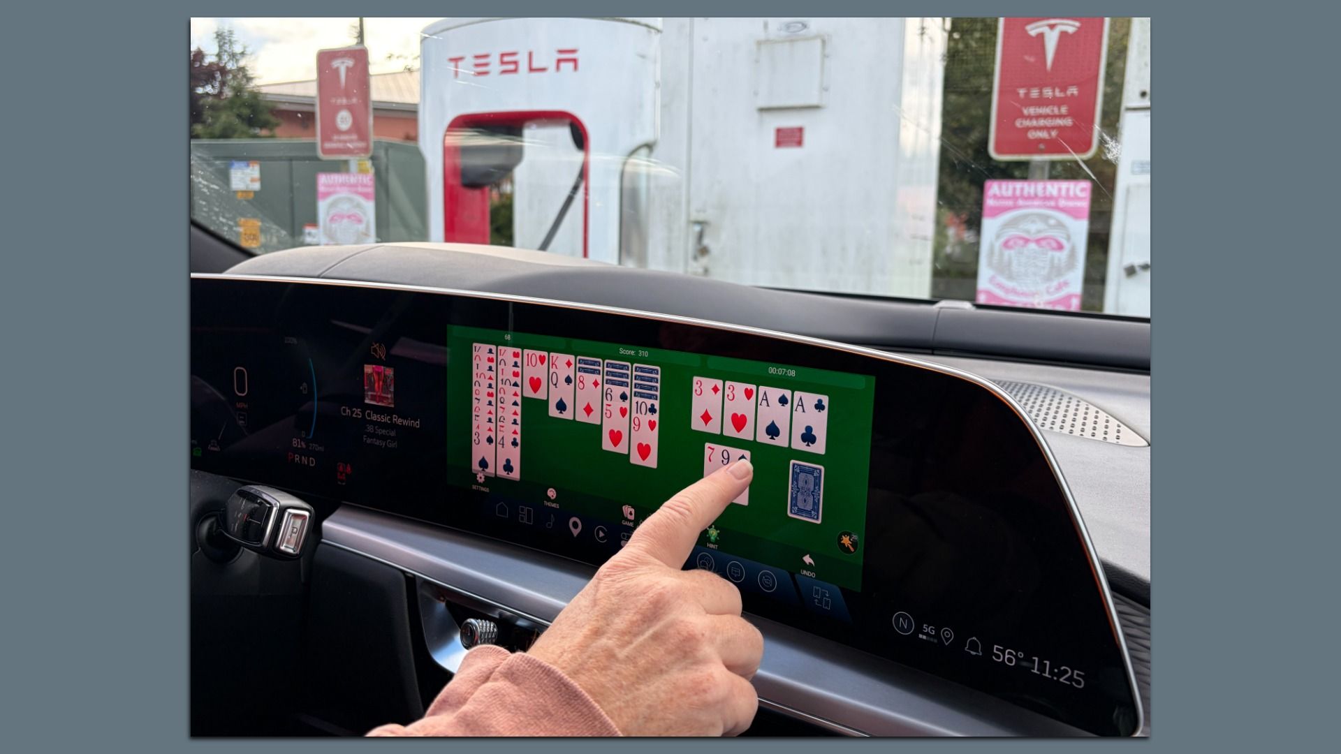 Close up of a car's large, horizontal touchscreen, displaying a Solitaire game on a green background. Outside, through the windshield a red Tesla Supercharger and signs are visible.