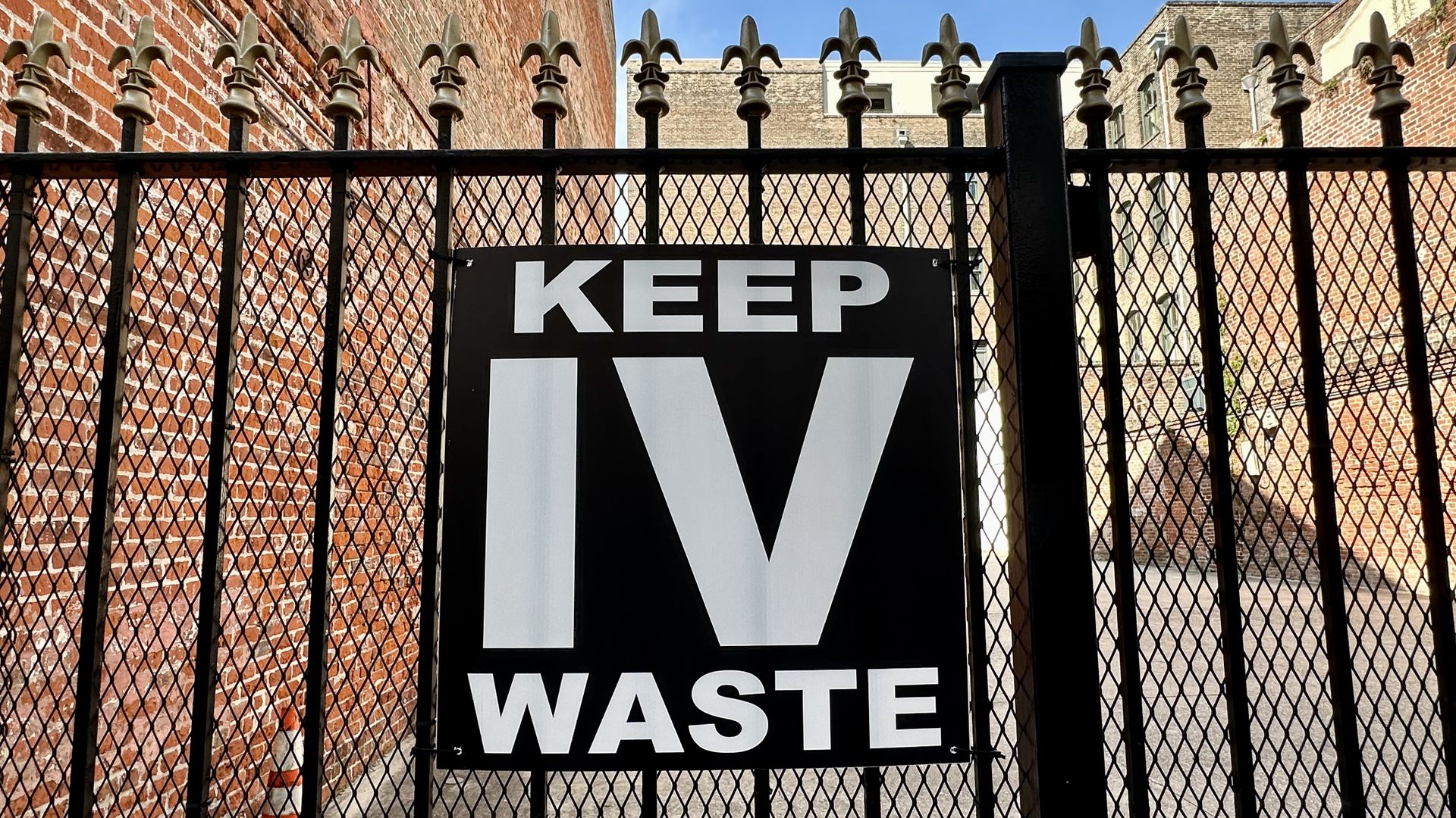 Black metal fence with pointed fleur-de-lis tops and a black sign reading KEEP IV WASTE in bold white letters, red brick walls on sides, clear blue sky above.