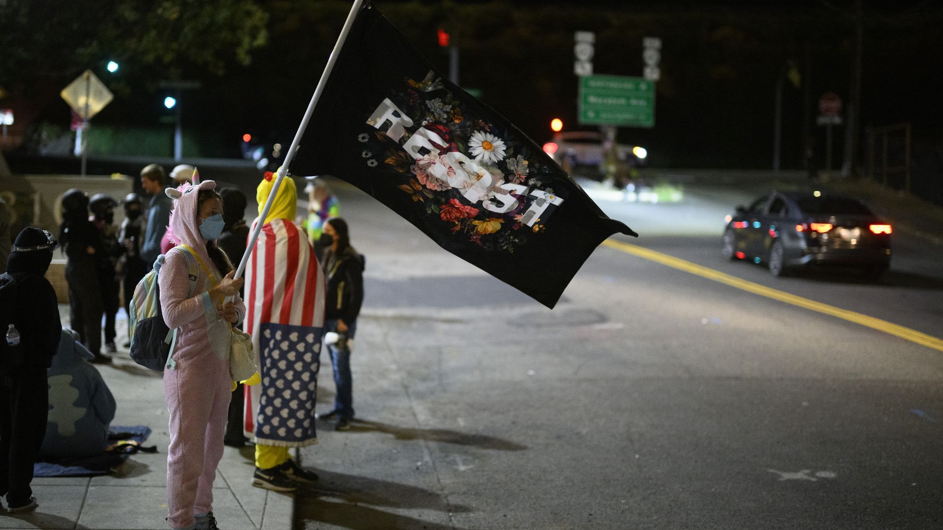 A person in a pink unicorn costume and mask holds a black flag with floral design and the word "Resist" at night, standing near people wrapped in American flag and other costumes by a street.