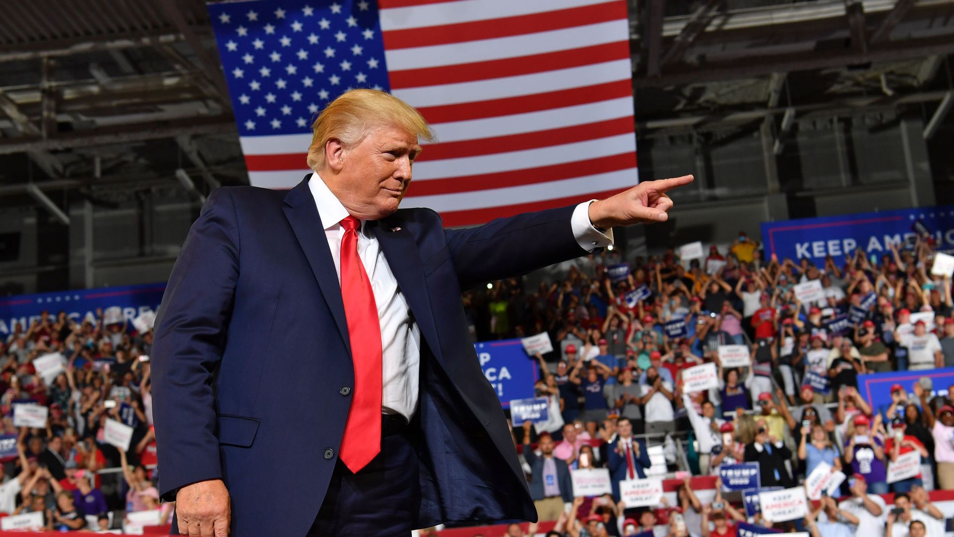 Trump stands on stage and points with an American flag behind him