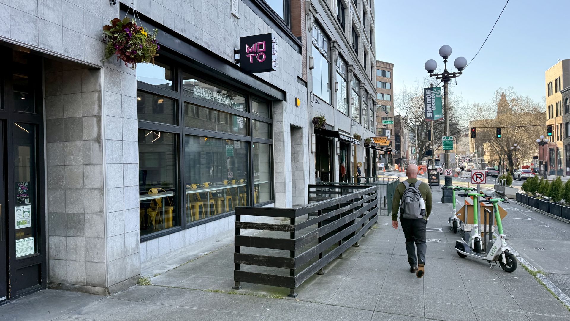 Man with a gray backpack walks along a city sidewalk past a gray stone building with large windows; a cafe with yellow chairs inside, hanging flowers, a black railing, and parked scooters along street.