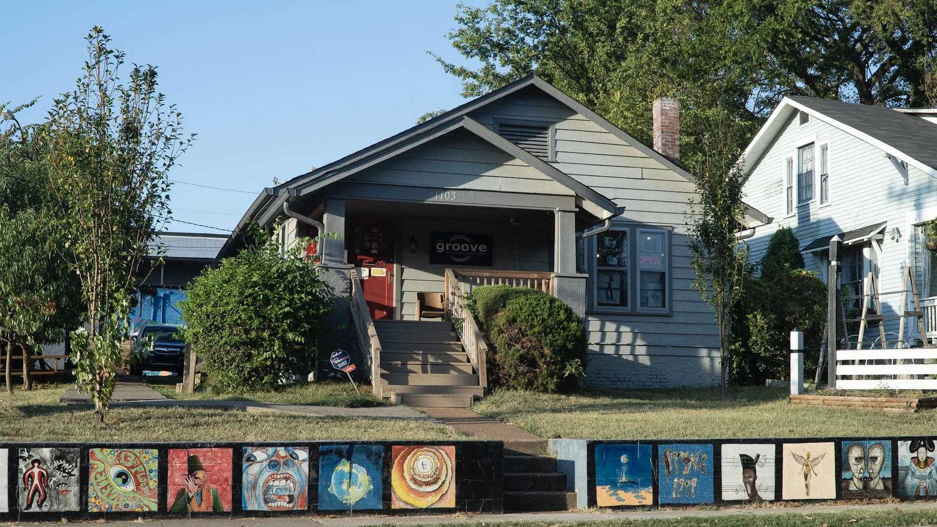 A gray house with a wooden porch and stairs, sign reading "groove," surrounded by green bushes and trees, and a mural of colorful artistic paintings on a low wall in front.