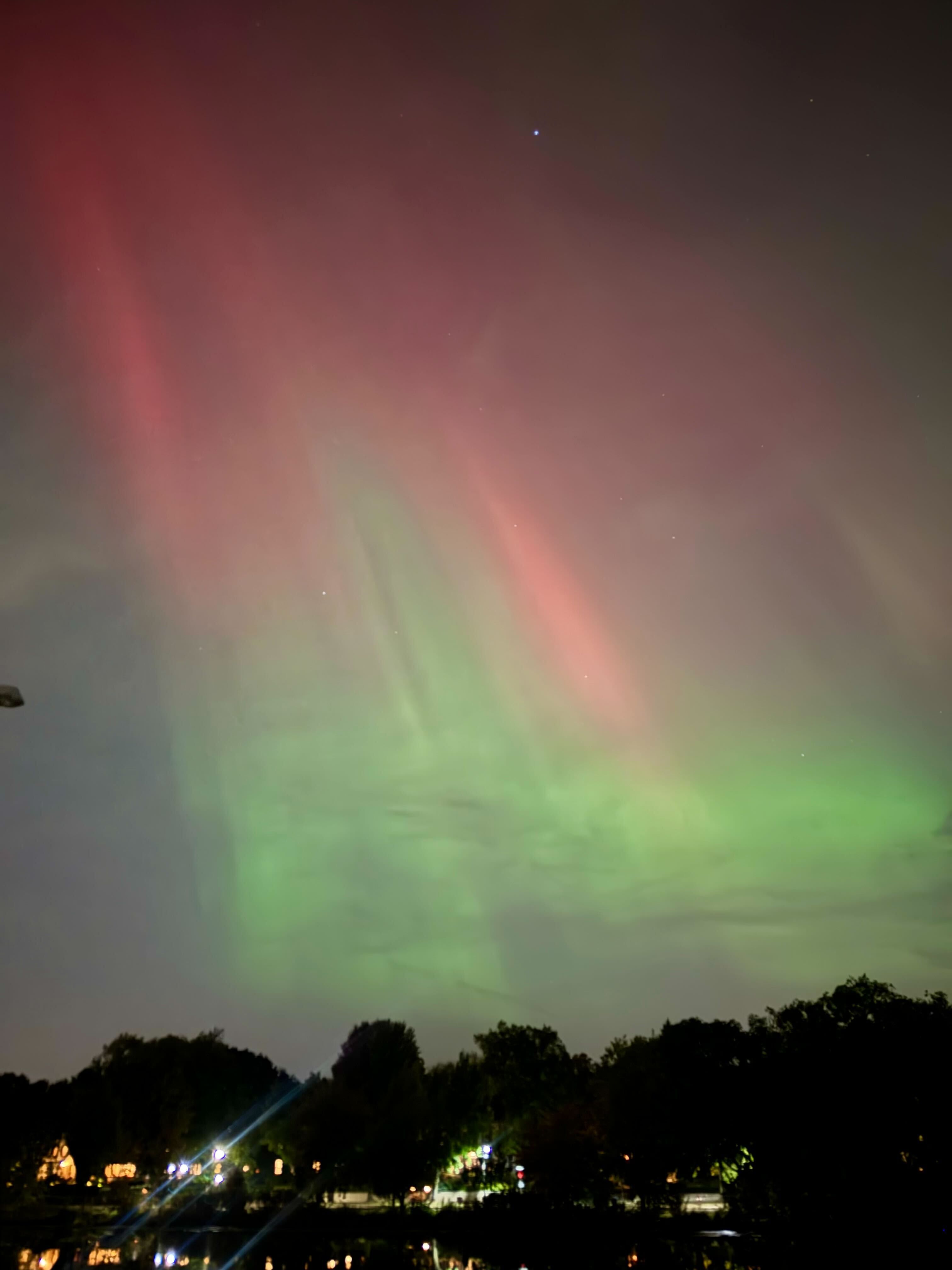 An aurora colored pink and green over trees and buildings with bright lights on the Lake of the Isles, about a mile from downtown Minneapolis.