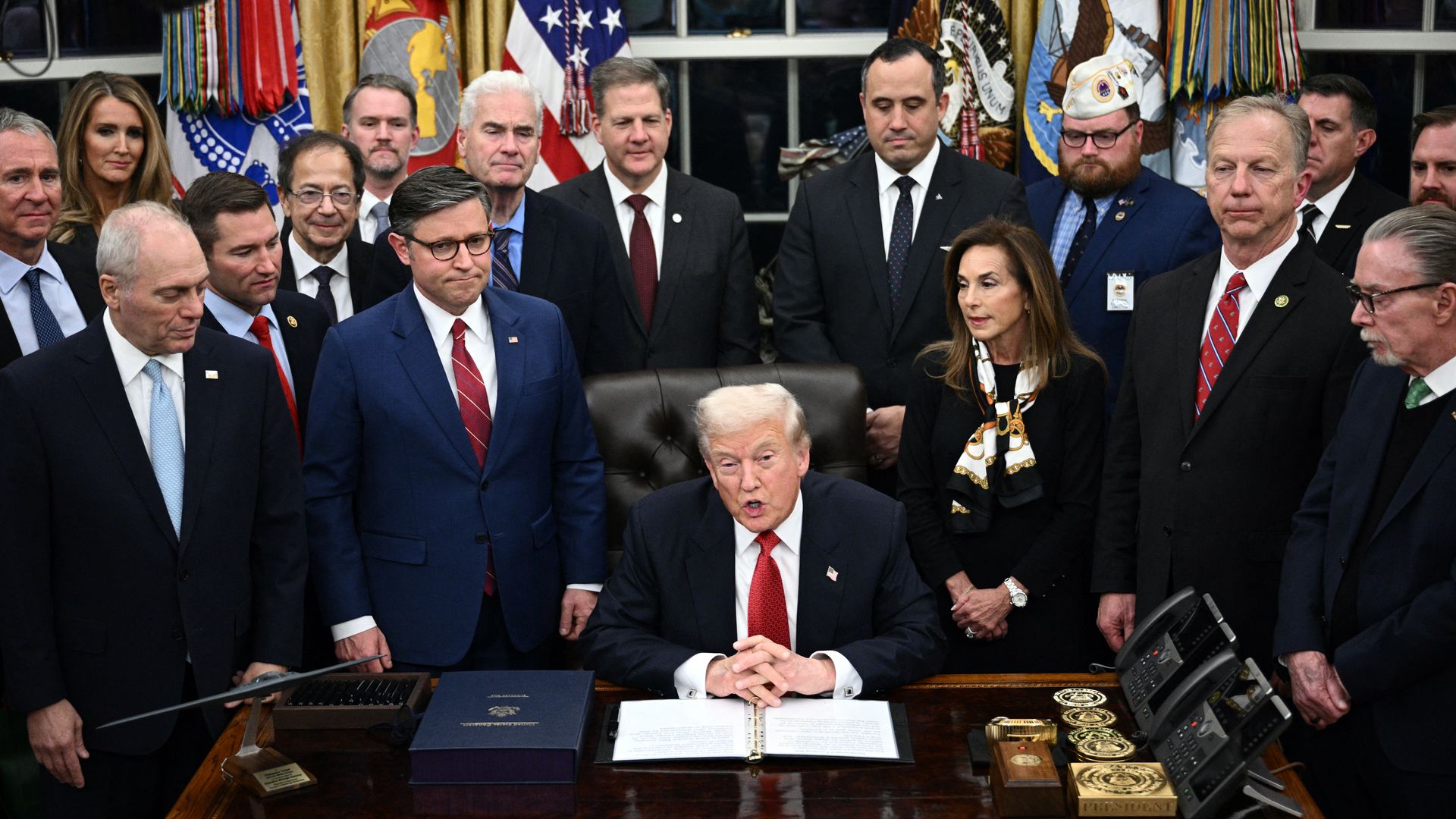 President Donald Trump signing a document at the Resolute Desk in the Oval Office, surrounded by a group of formally dressed men and women, with U.S. flags in the background.
