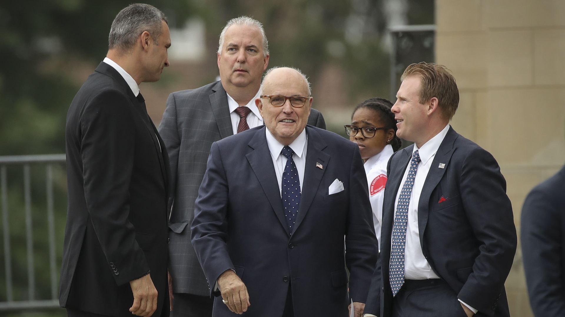 Rudy Giuliani walking toward the camera in a navy suit and brown glasses surrounded by three other white men in blue suits in discussion