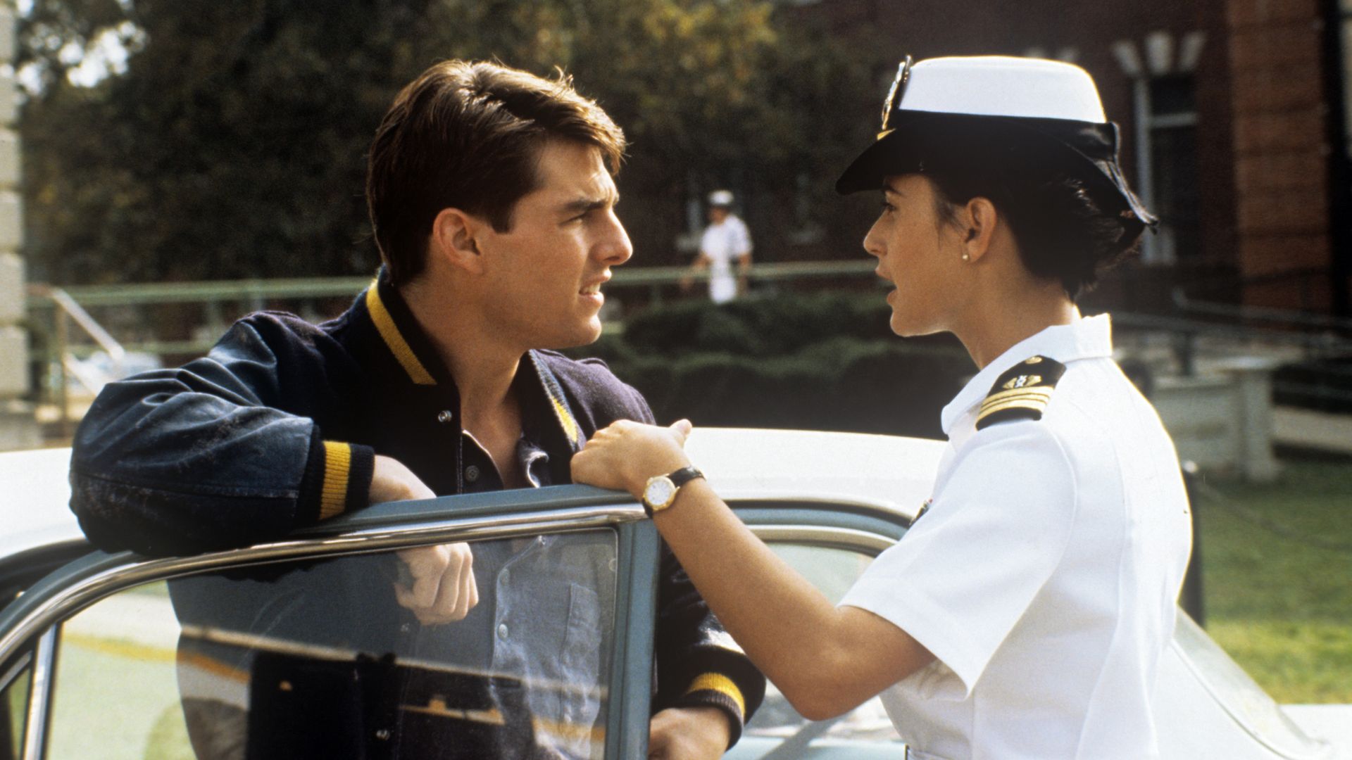 A man in a dark blue jacket talks to a woman in a white naval uniform and hat by a car door, with a brick building and greenery in the background.