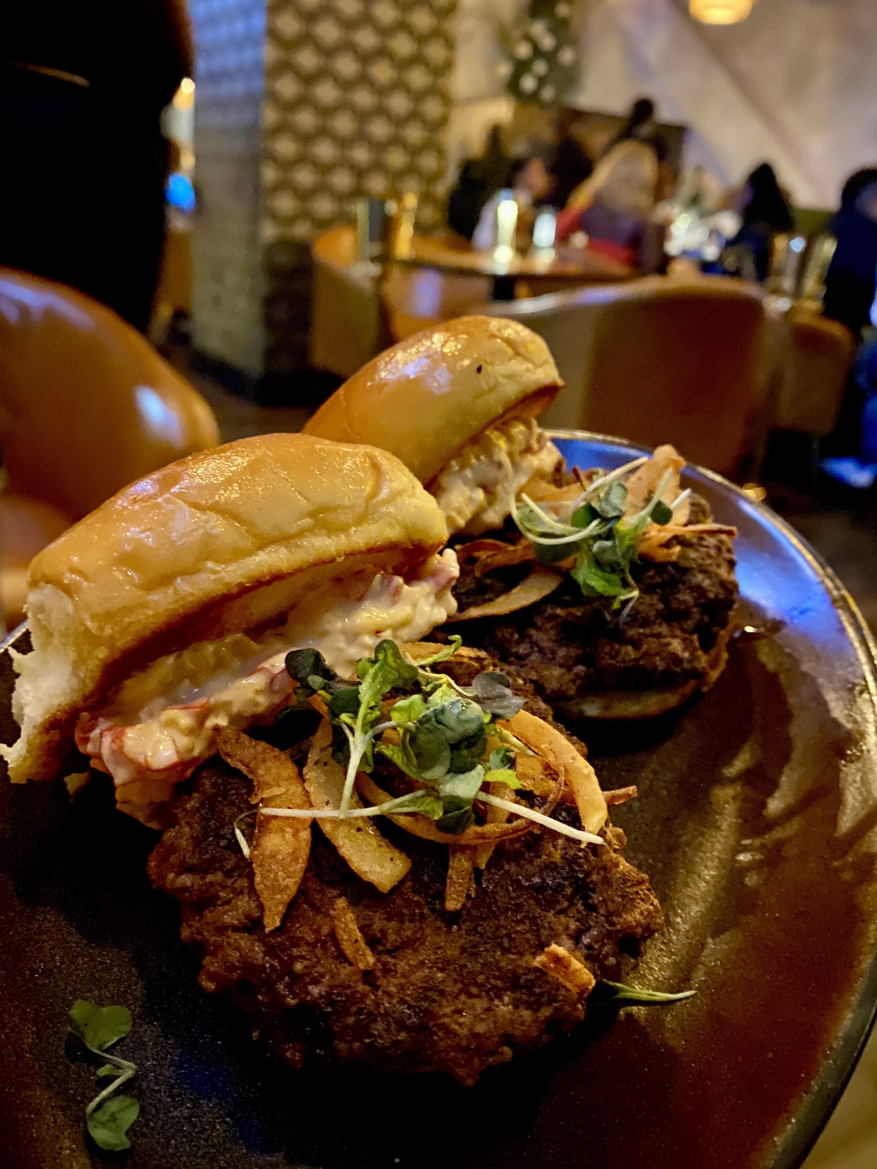 Two gourmet sliders with toasted buns, topped with crispy fried onions, microgreens, and creamy sauce on a dark plate in a cozy restaurant setting.