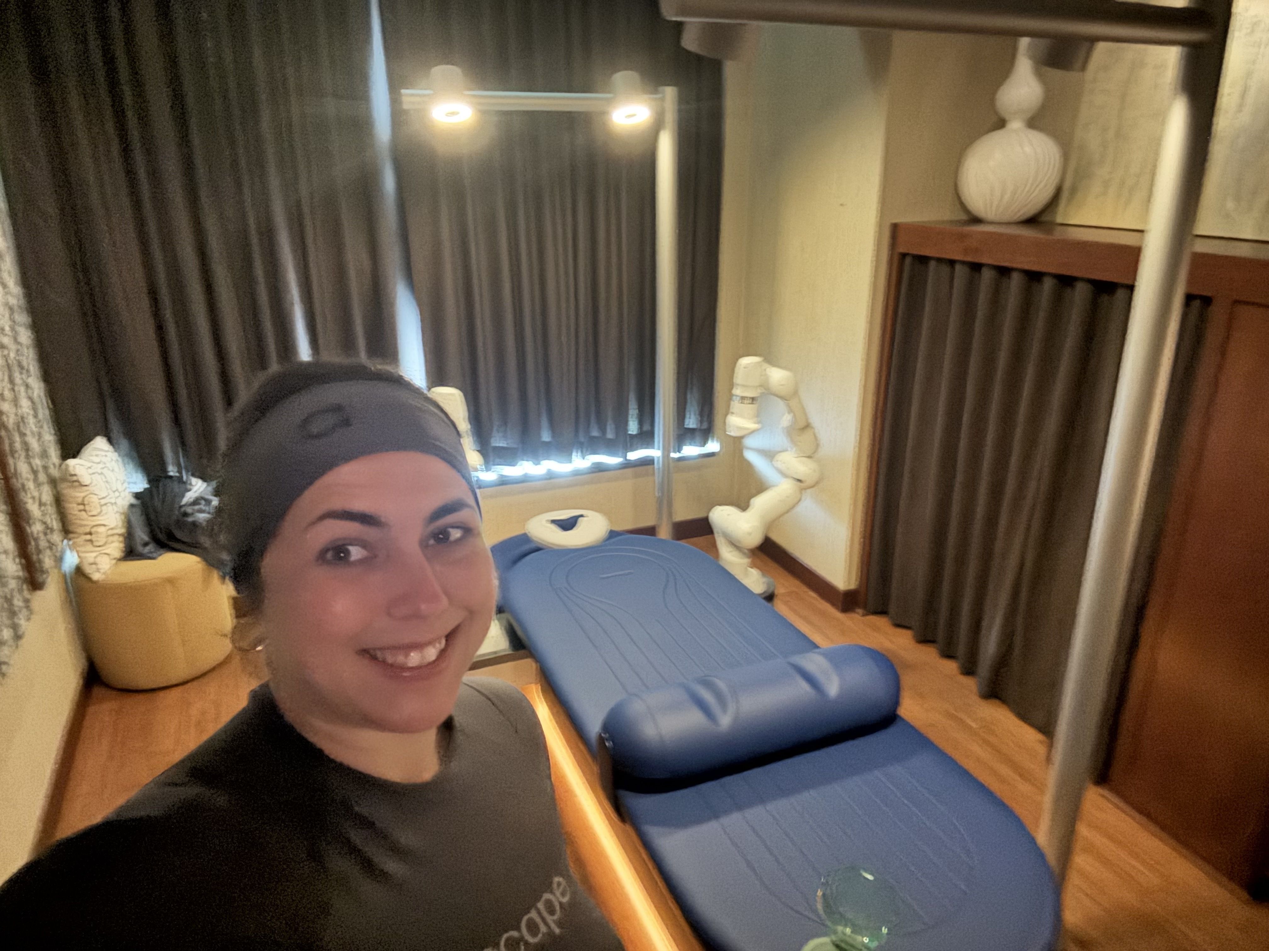 Smiling woman in black headband and shirt takes selfie in a room with wooden floor, dark curtains, a blue massage table, and a white decorative sculpture in corner.