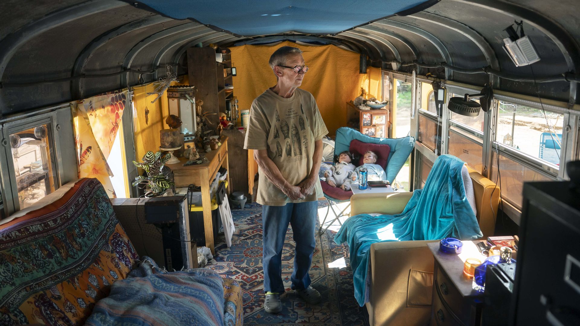 An elderly person stands inside a cozy, decorated bus interior with colorful blankets, a patterned rug, toys, and furniture including chairs and a dresser under a blue ceiling.