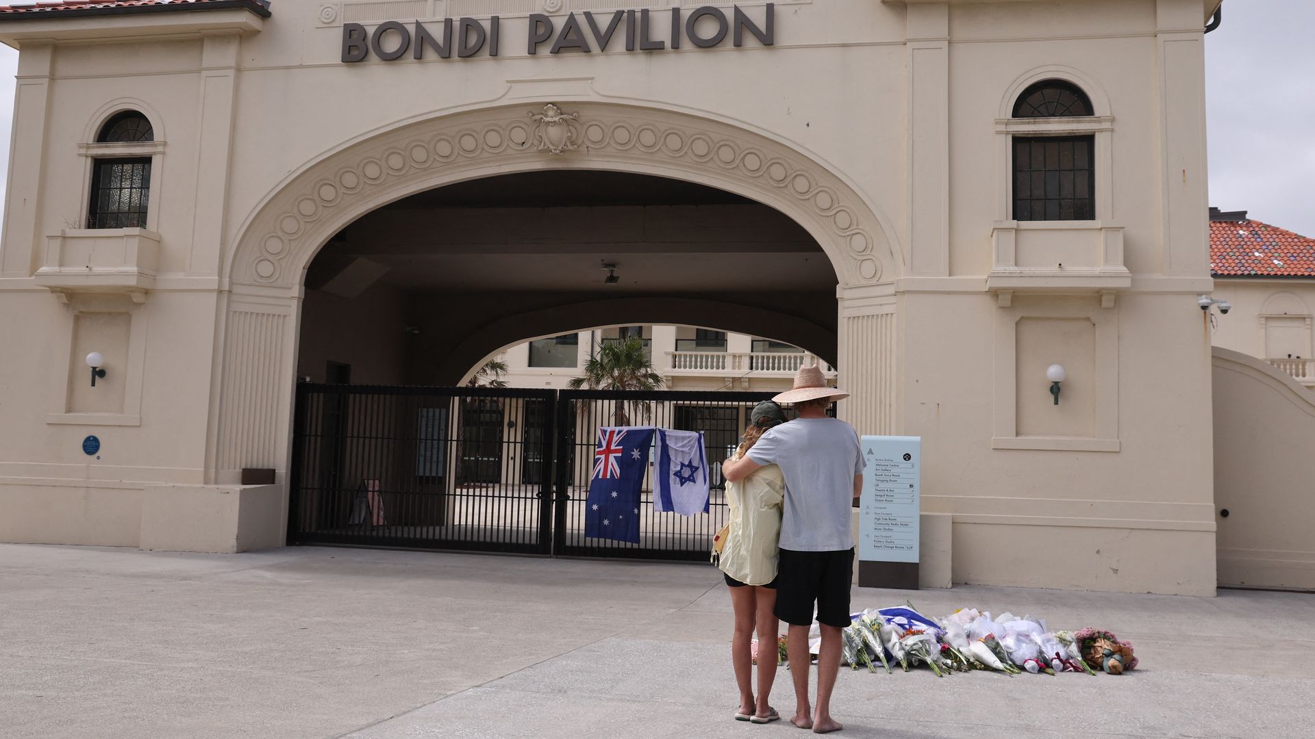 The back of a man in a large straw hat, light blue shirt, black shorts and flip flops holding a woman in a black baseball cap, yellow t-shirt, small, black shorts and flip flops as they stand before a makeshift floral memorial to victims of a mass shooting at Bondi Beach in Sydney.
