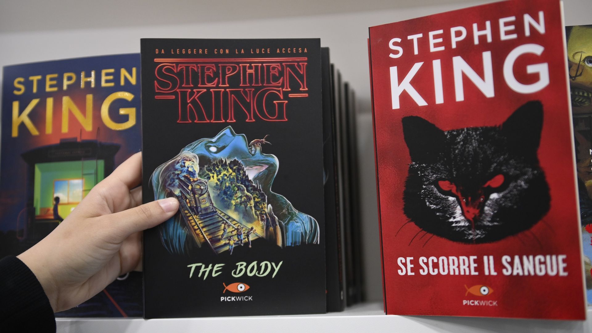 Hand holds a black Stephen King book titled "The Body" with red lettering; blue cover on the left and a large red Stephen King cover on the right on a bookshelf.