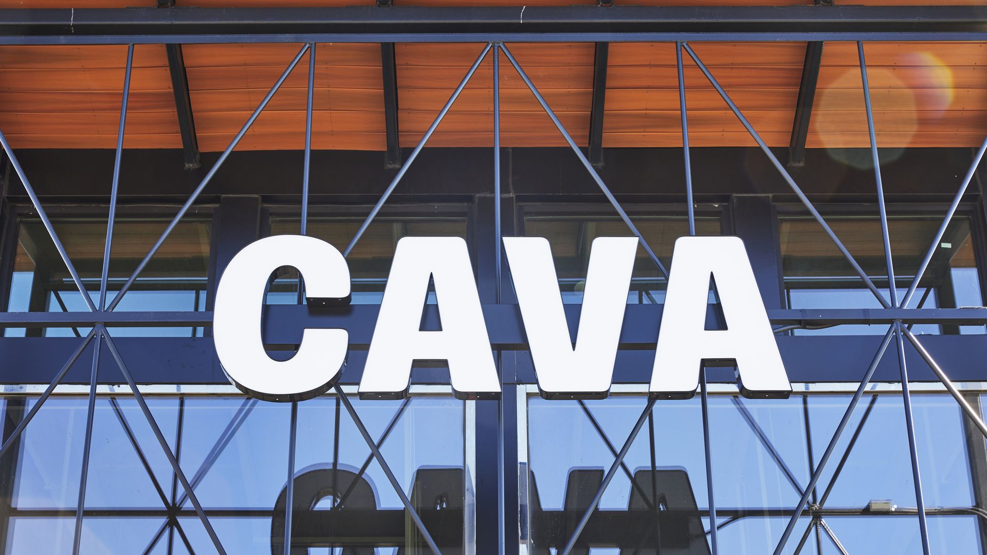 White letters spelling "CAVA" on a building facade with large glass windows and black metal framework, reflecting the text against a blue sky and wooden ceiling background.