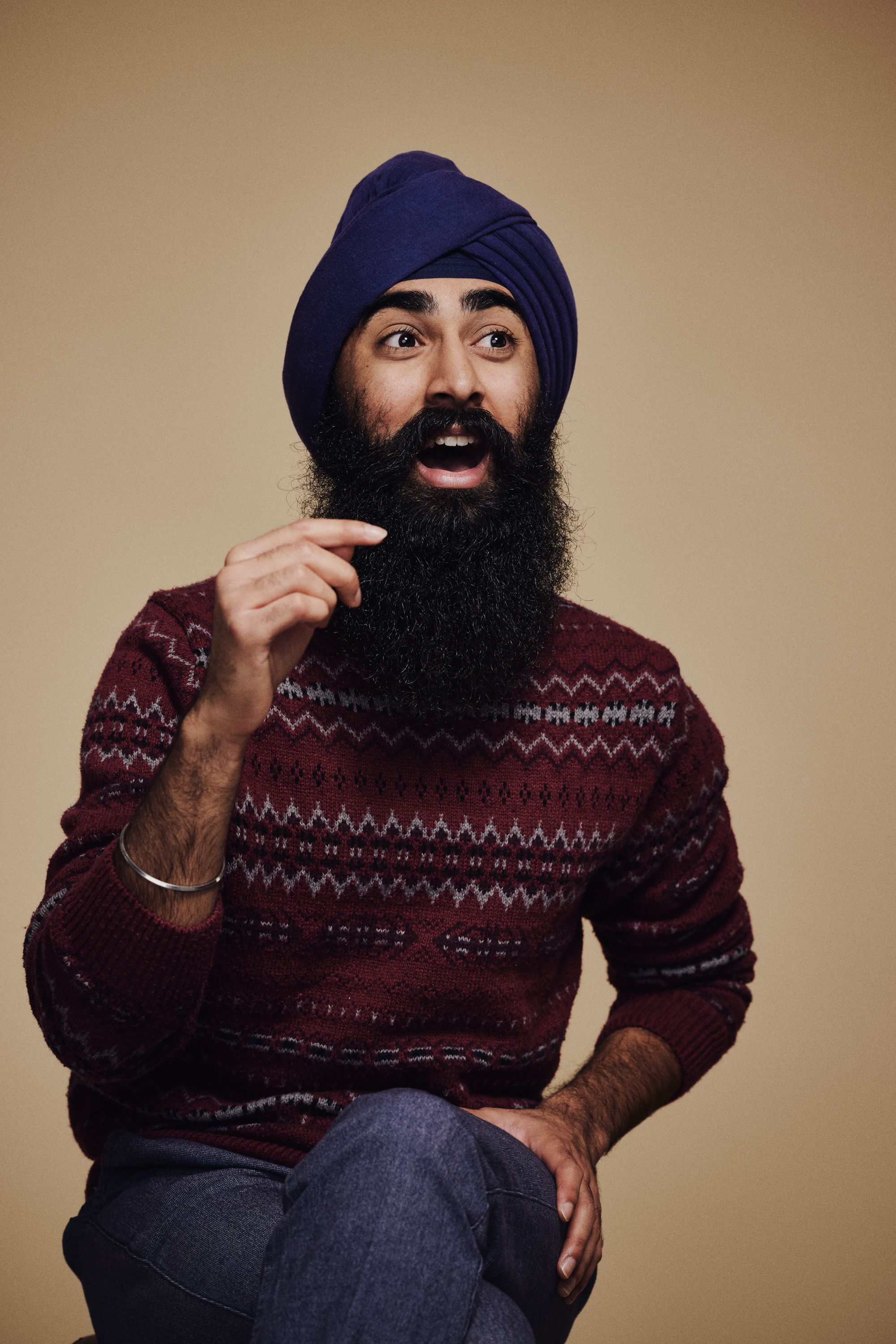 A man with a blue turban and full black beard sits against a beige background, wearing a maroon patterned sweater, gesturing with his right hand as he speaks.