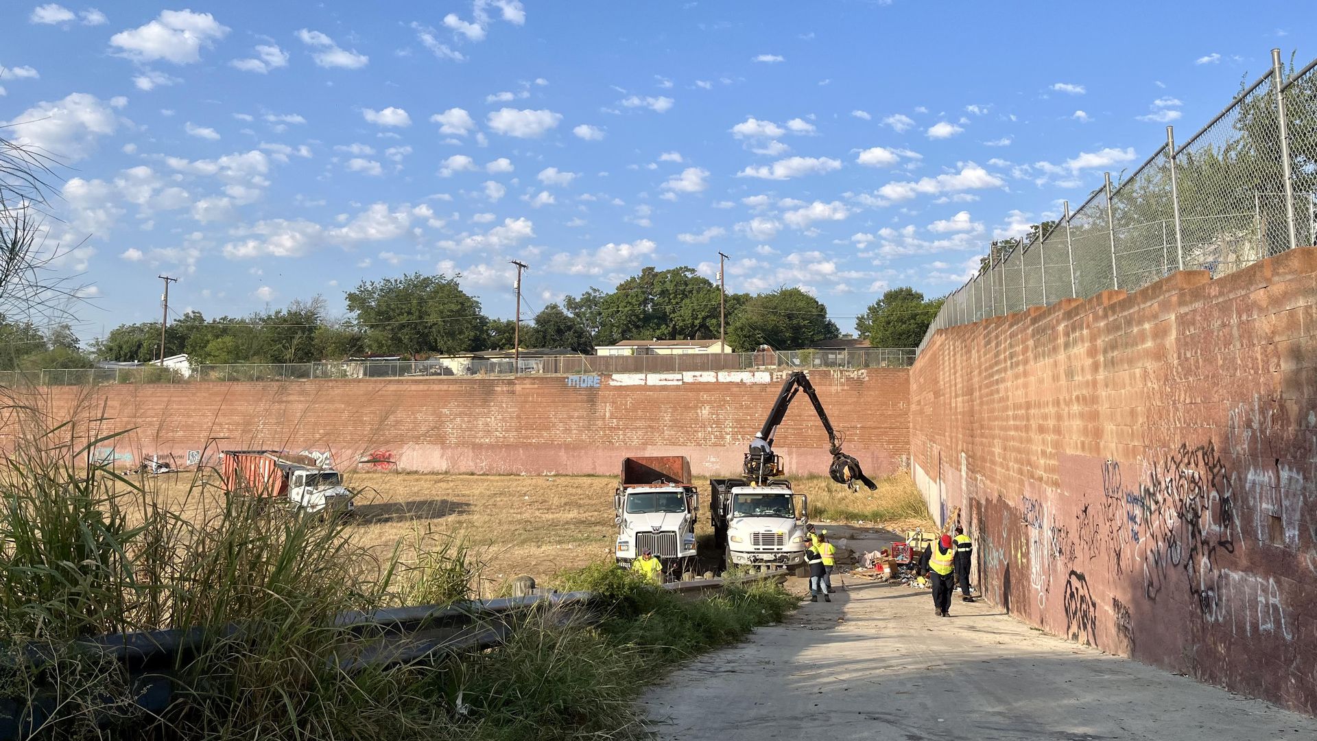 Dump trucks pick up trash as cleanup crews walk a field surrounded by tall brick walls under an open blue sky.