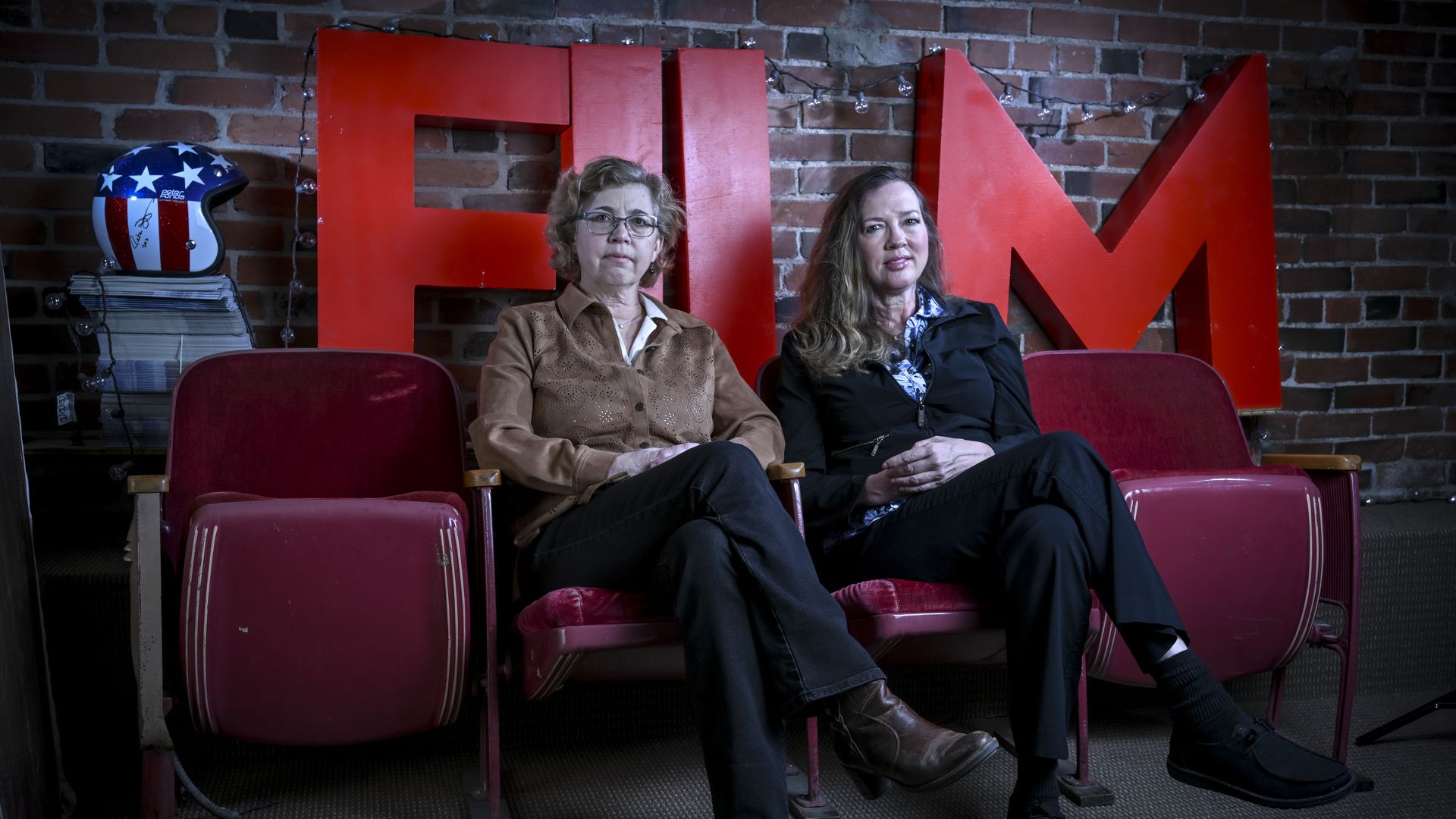 Two women sit on red vintage theater seats against a brick wall, with large red letters behind them. On the left, a blue helmet with white stars sits on a stack of papers.