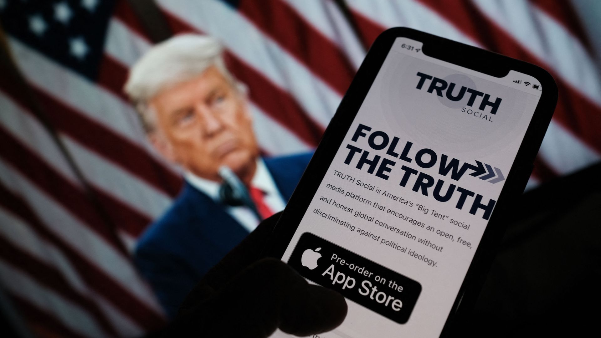 A person checking the app store on a smartphone for "Truth Social", with a photo of ex-president Trump on a computer screen in the background, in Los Angeles, October 20