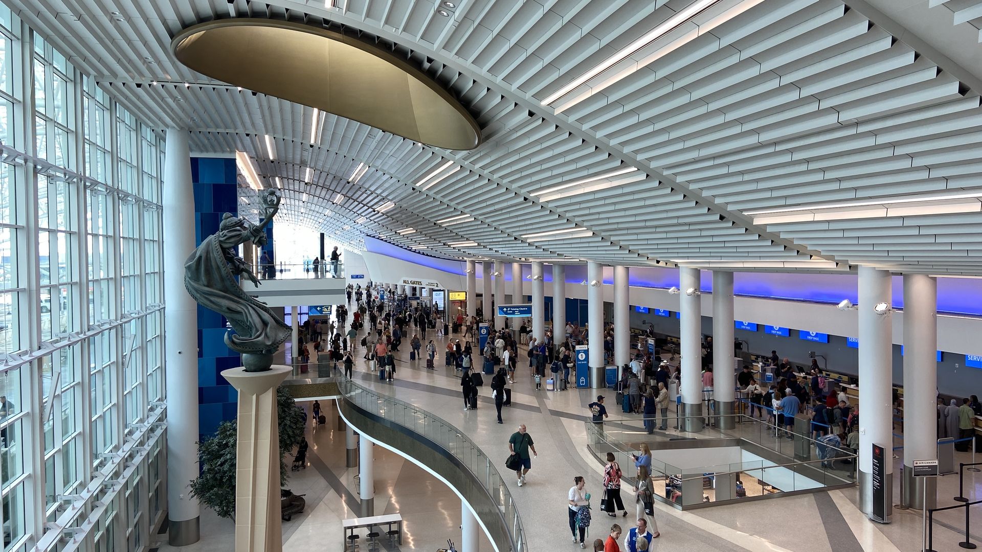 Inside a bright airport terminal: a curved white slatted ceiling, glass walls, a statue on a pedestal in the foreground, and crowds of travelers at blue-lit check-in counters.