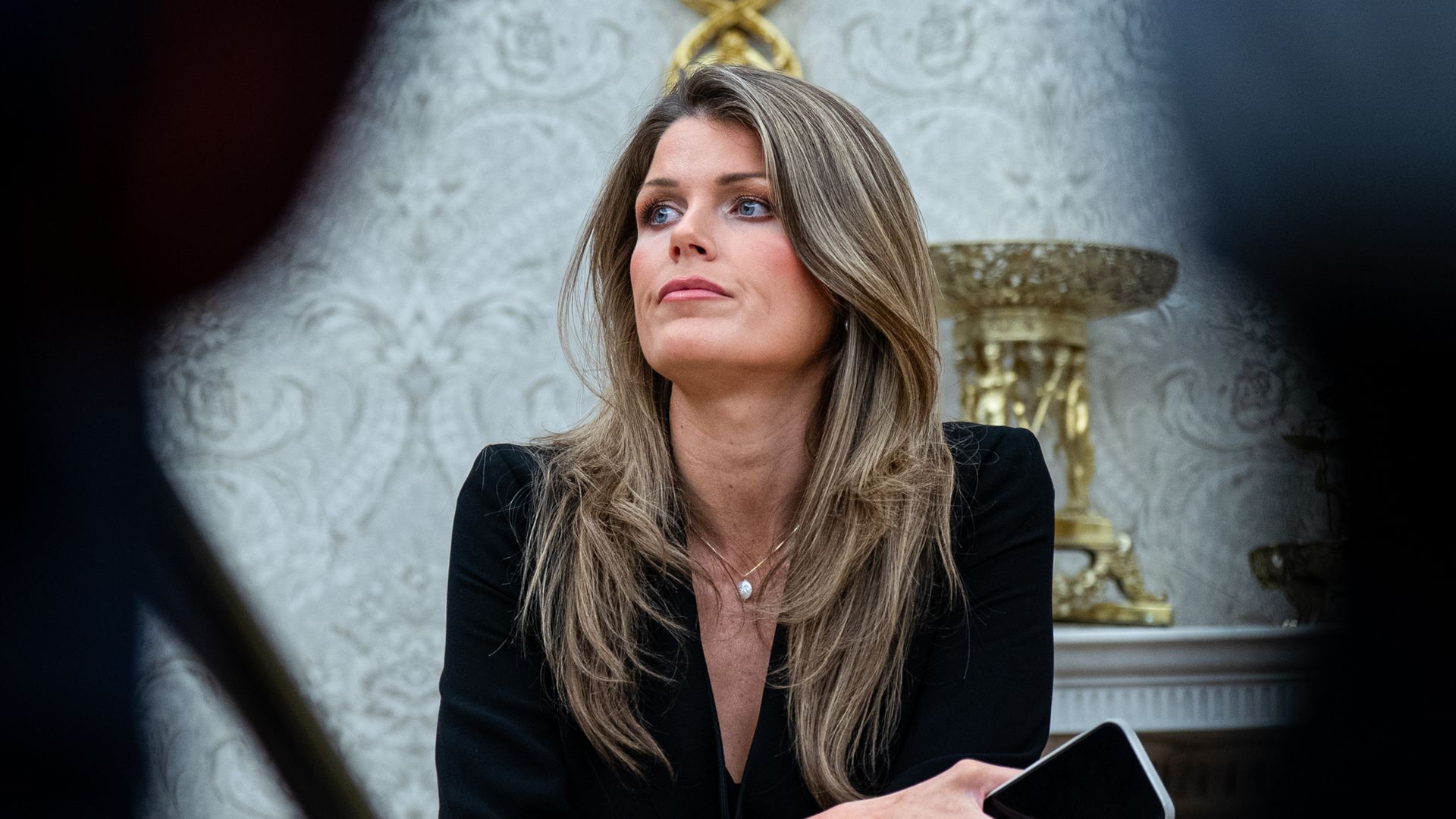 A woman with long light brown hair, attorney Lindsey Halligan, wearing a black outfit, holds a smartphone and looks to the side. She is seated in an ornate room with a gold decorative element on the wall behind her.