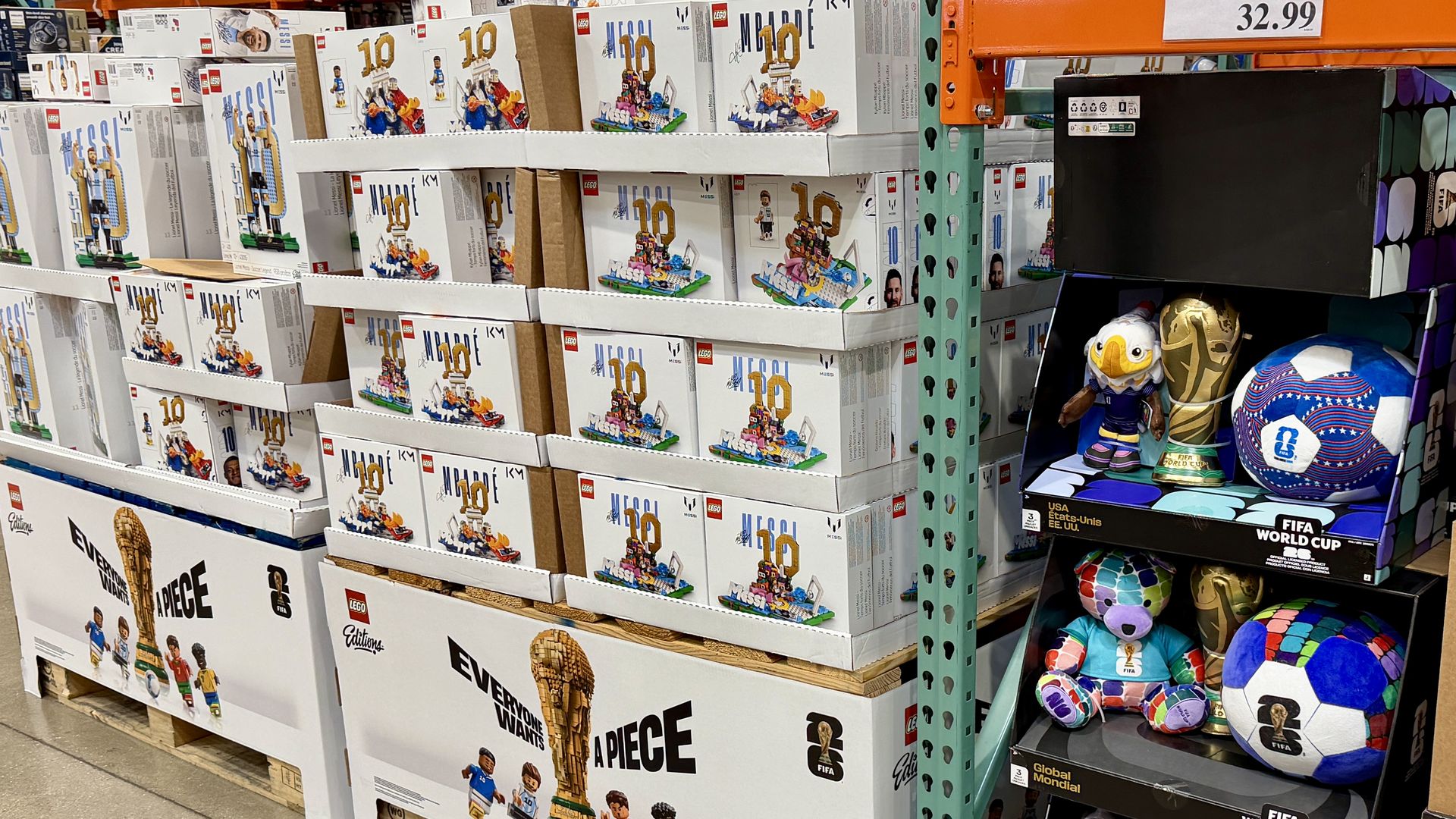Store aisle full of LEGO boxes stacked on shelves, featuring minifigures and the LEGO logo; right side has FIFA World Cup plush toys and a boxed soccer ball set, price tag $32.99.