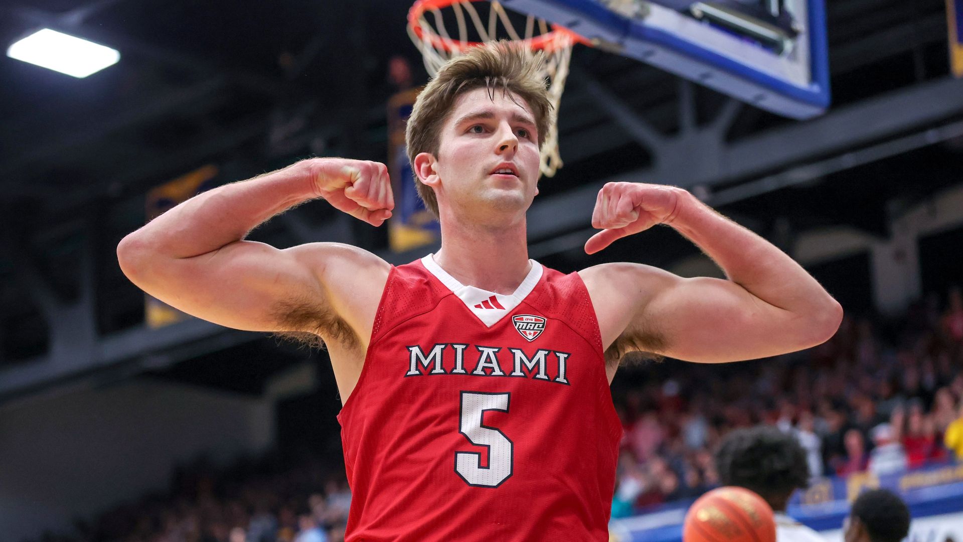 Basketball player in red Miami jersey number 5 flexes muscles under a basketball hoop during a game, with blurred players and crowd in the background.