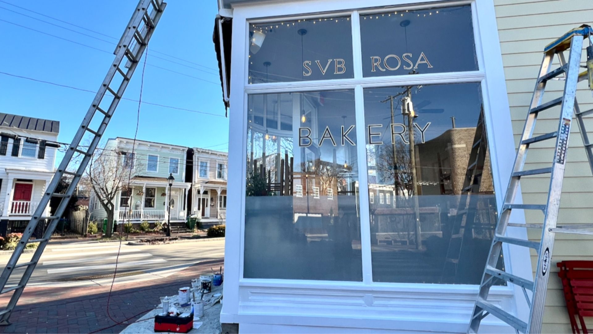 Exterior of SVB Rosa Bakery with large glass windows, white trim, ladders on both sides, and paint supplies on the sidewalk under a clear blue sky in a residential area.