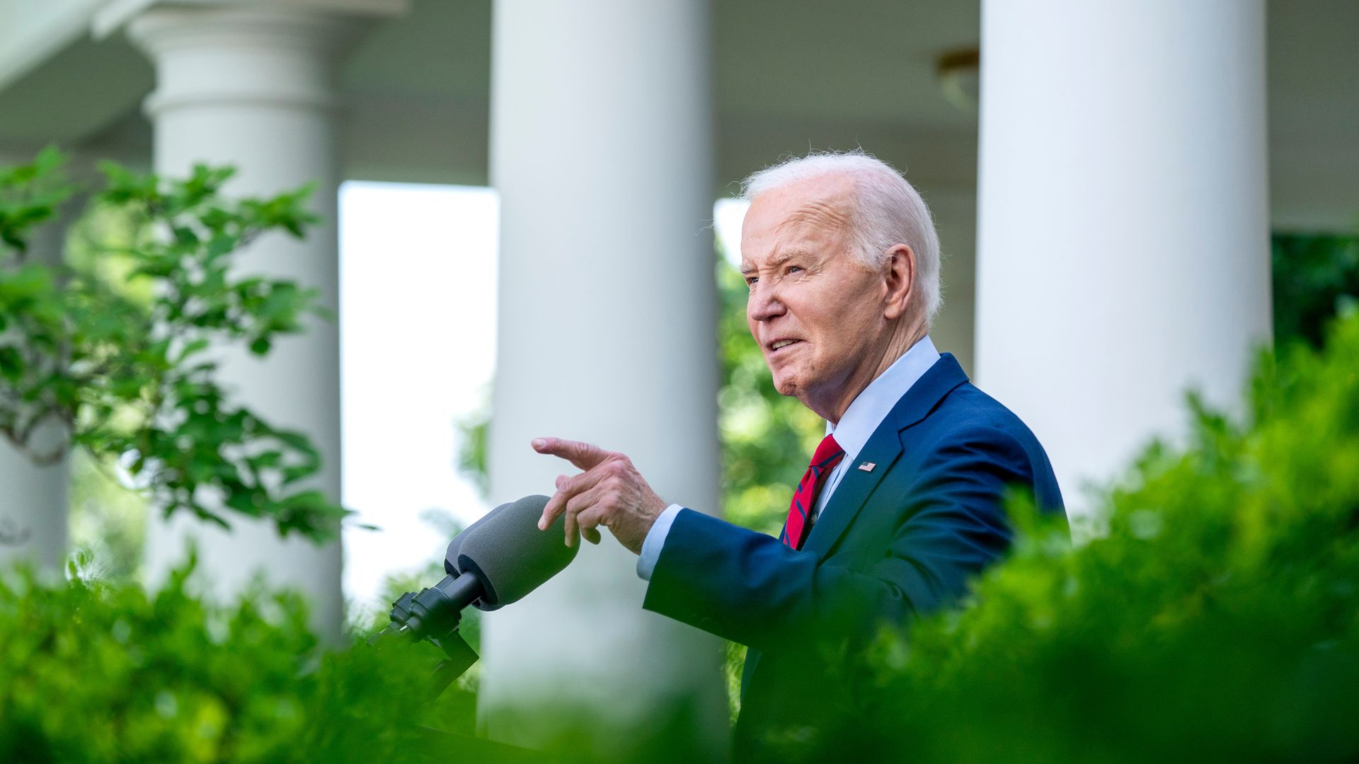 President Biden, in a blue suit and red tie, is seen speaking into a microphone outside the White House with bushes in the foreground.