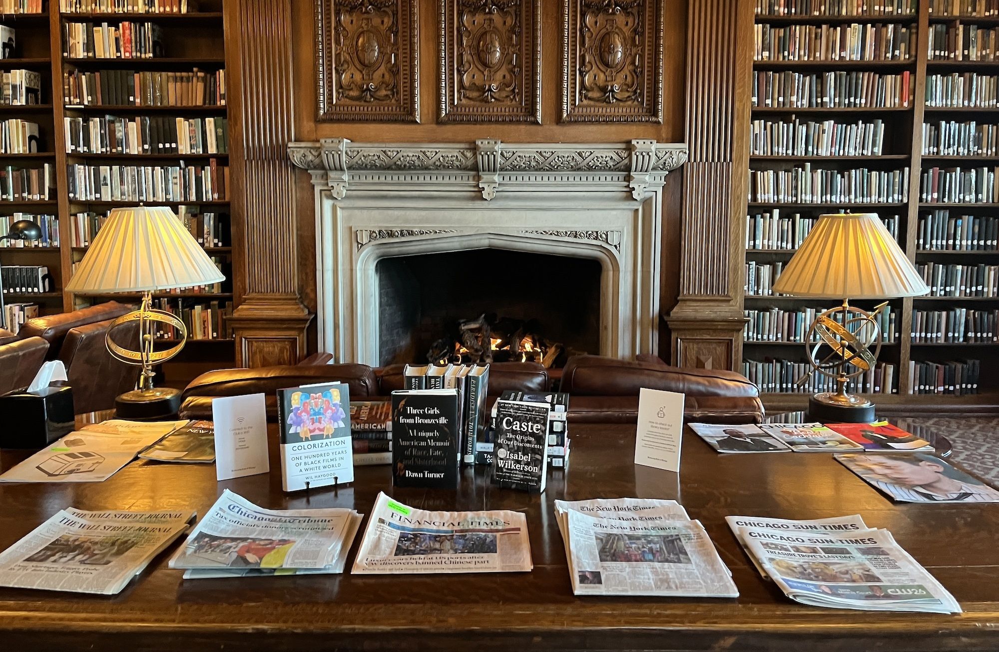 A fireplace in a large library with newspapers and books on a table.
