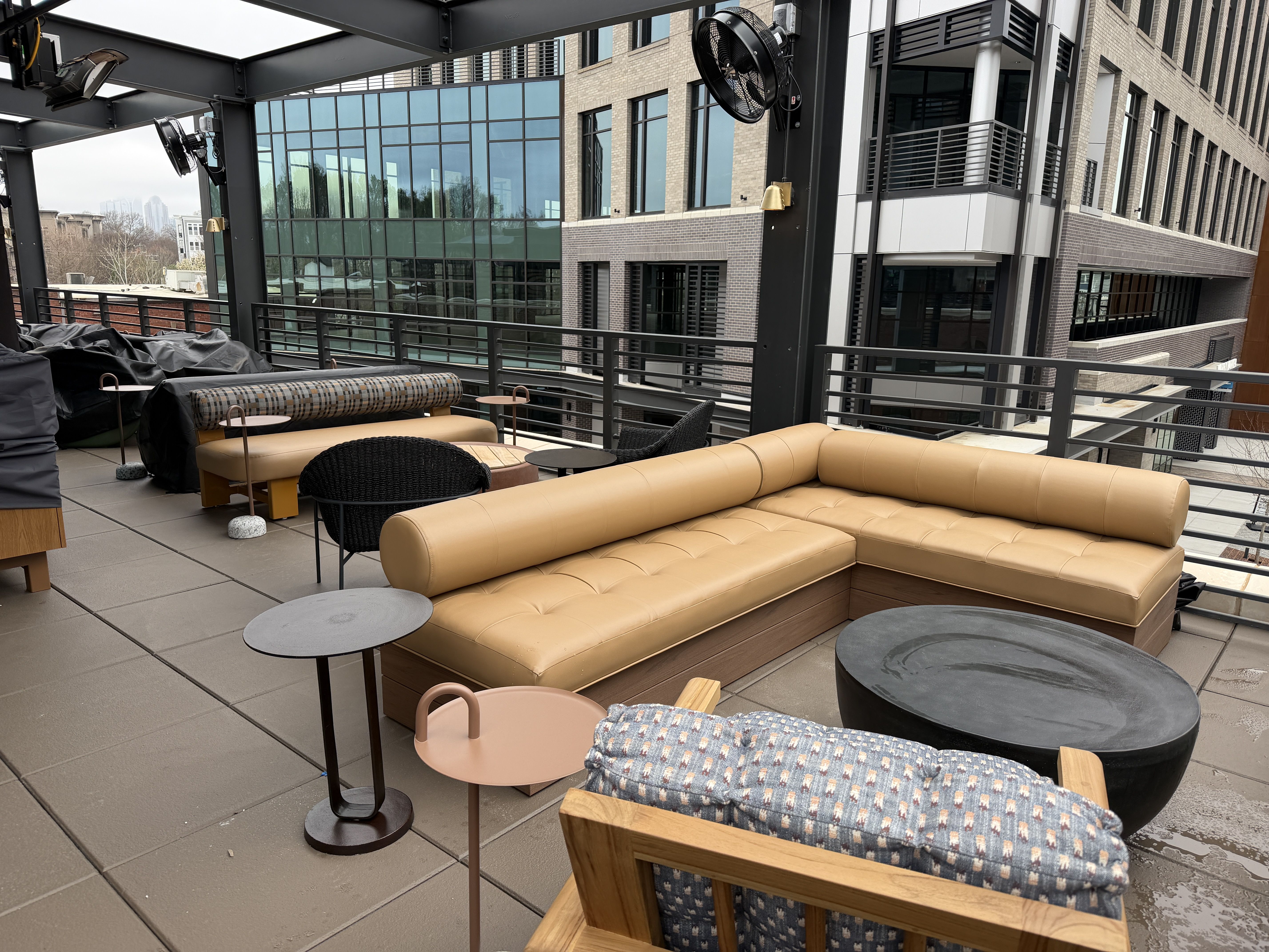 Outdoor patio seating area with tan leather corner sofa, black and wood chairs, small round tables, and large circular black fire pit, surrounded by modern office buildings under a metal pergola.