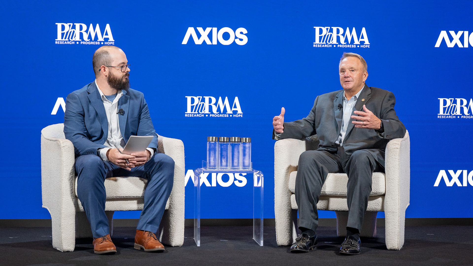 Two men in suits sit on beige chairs on a blue stage, speaking in a panel. The backdrop shows PhRMA and Axios logos; a clear acrylic table with water bottles sits between them.