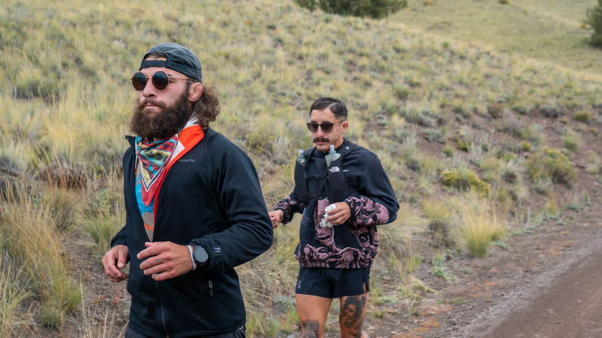 Two hikers walk a dirt trail across a dry, grassy hillside. Foreground: bearded man with a backward cap, sunglasses, and colorful scarf; behind him: tattooed man with a backpack.