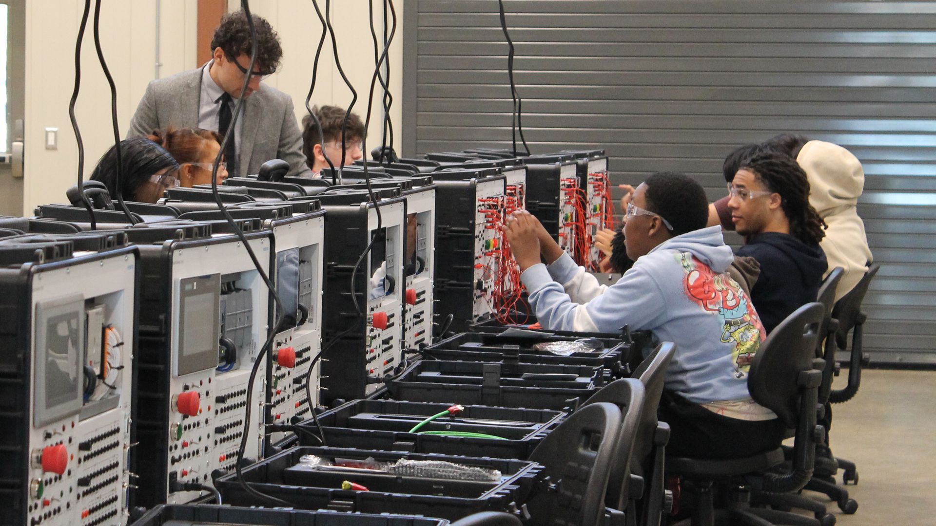 Students working with wiring setups on black electronic stations in a large room, guided by two instructors. Ceiling-mounted plug connectors hang above each station.