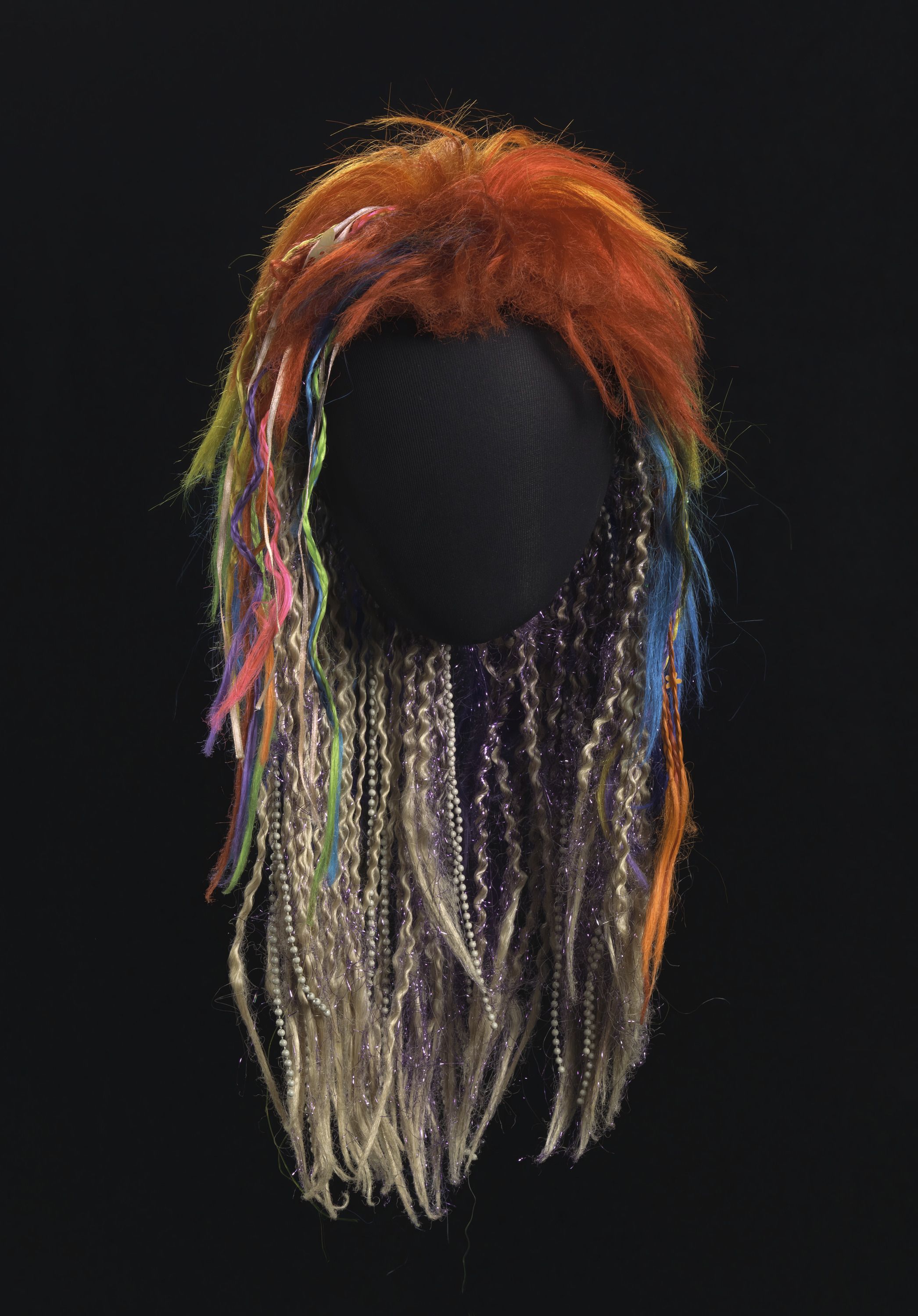 A multicolored wig, with blue, red, orange and green, sits on a mannequin head with a black background. 