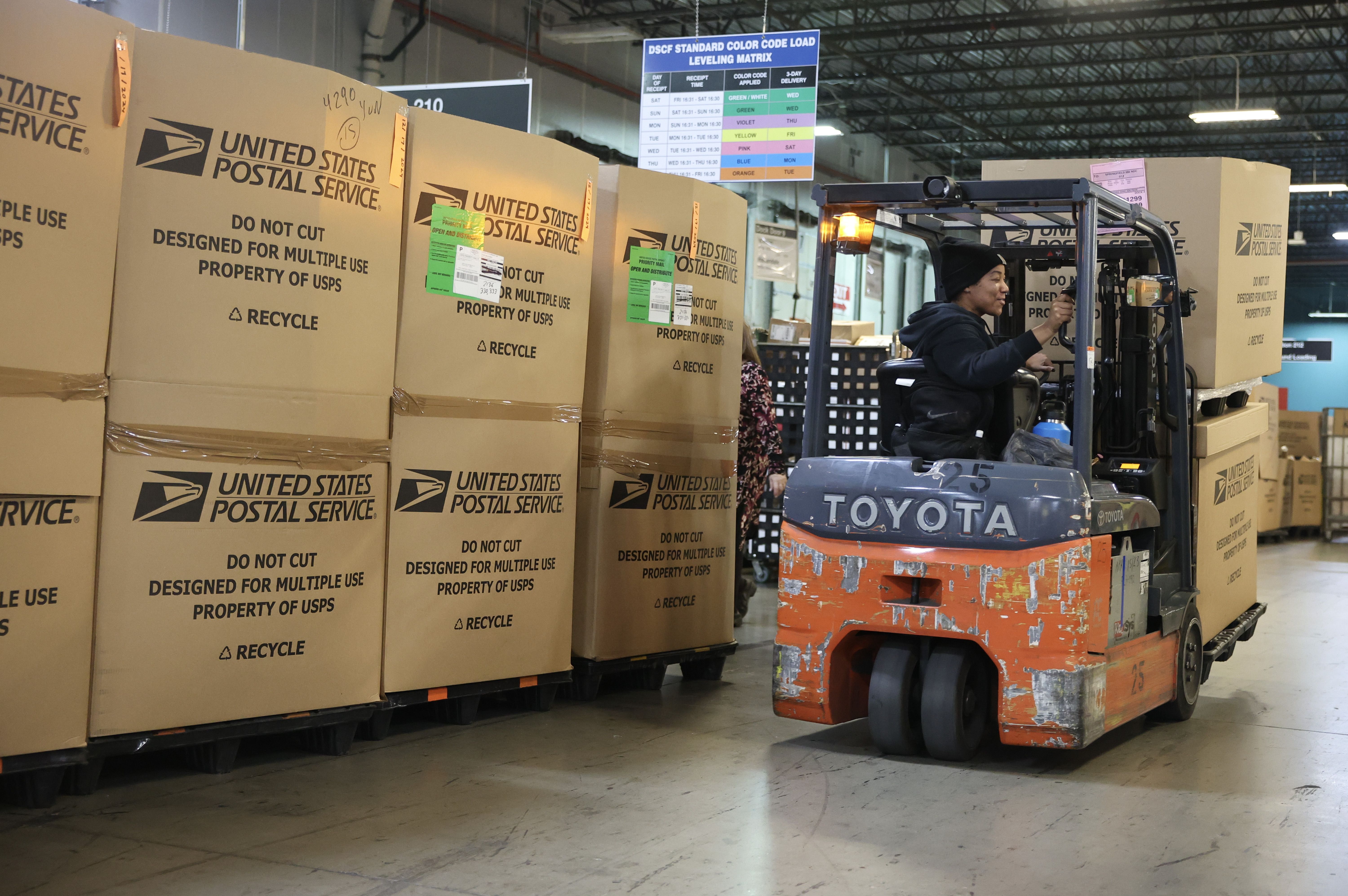 Worker operating an orange Toyota forklift moving large stacked USPS cardboard boxes inside a warehouse with a color-coded delivery sign visible in the background.