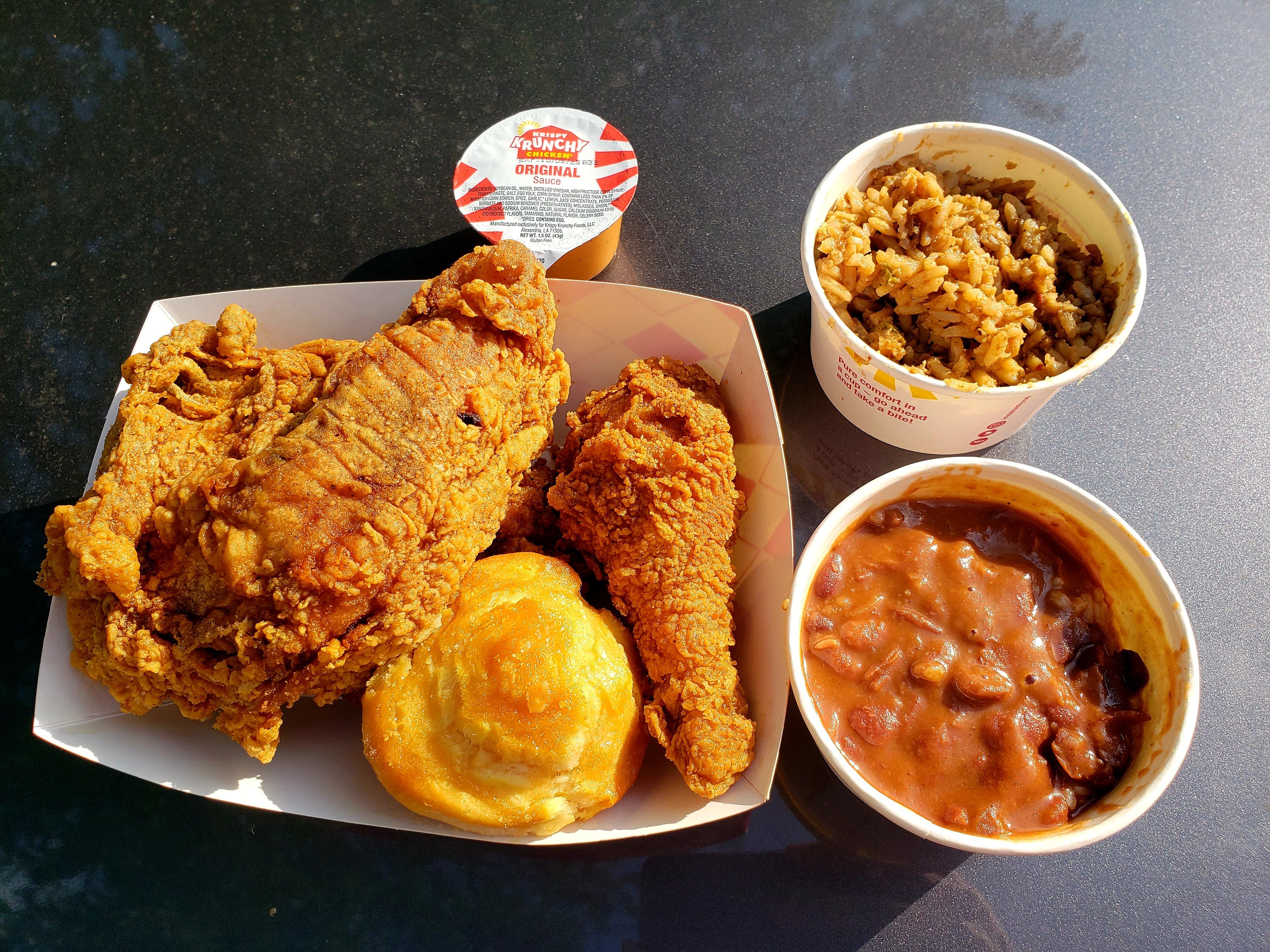Krispy Krunchy chicken in a paper tray with a biscuit, next to a cup of jambalaya and red beans and rice. 