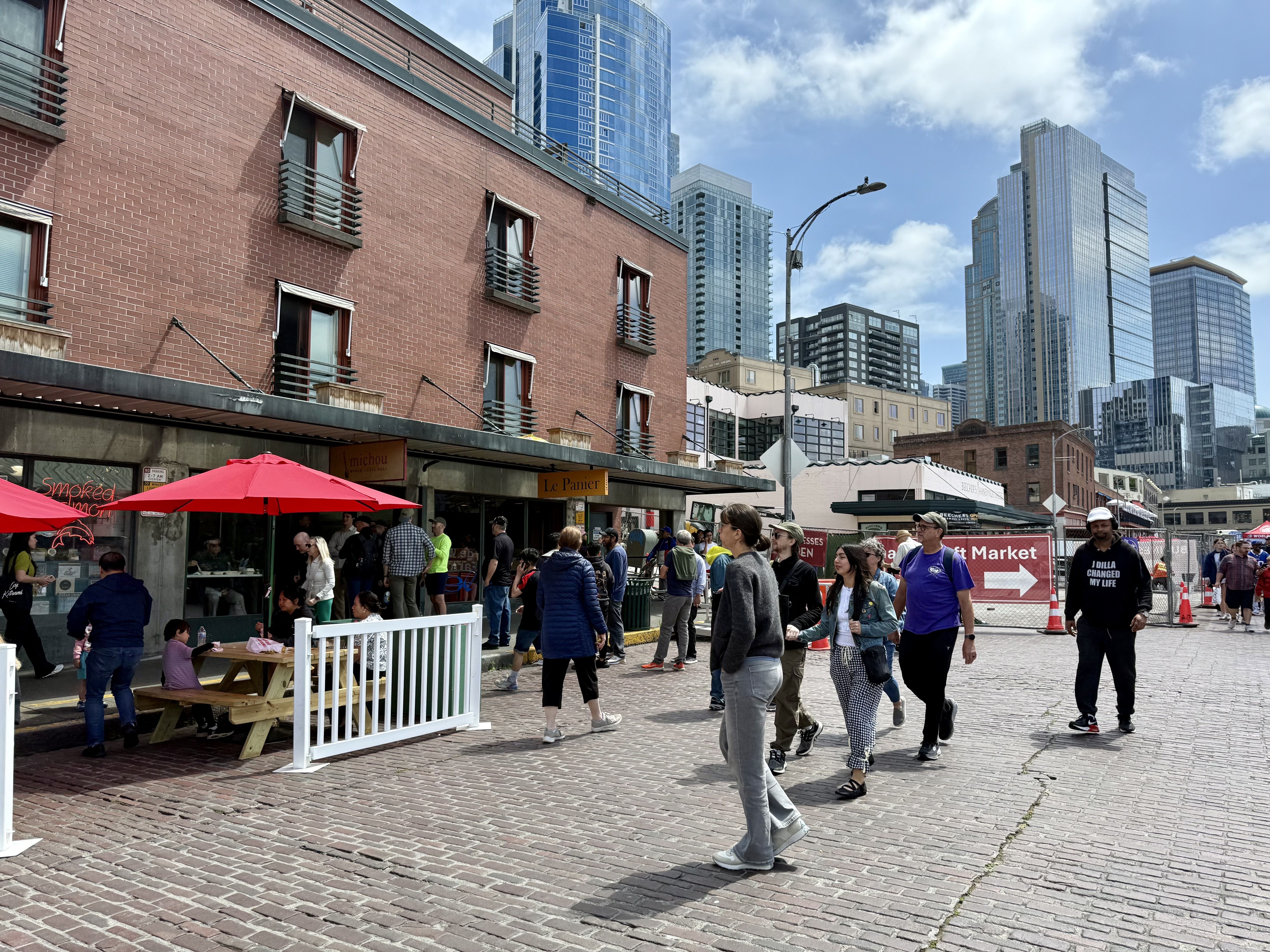 People walk along Pike Place in Pike Place Market with umbrellas over seating at left and a view of construction in the background.
