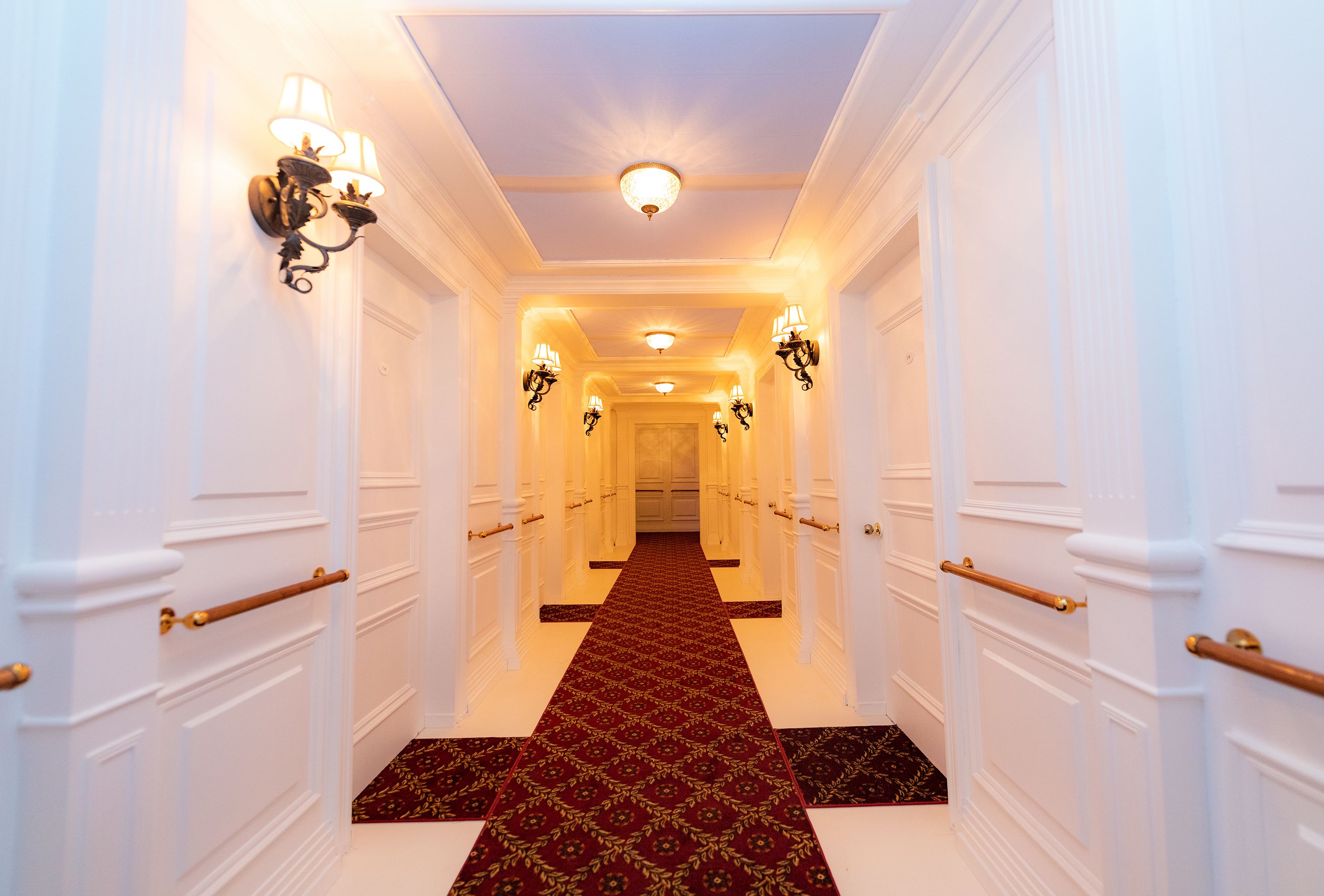 Bright hotel hallway with white paneled walls, doors lining both sides, ornate black wrought-iron wall lamps, brass railings, and a burgundy patterned runner carpet extending down the corridor.
