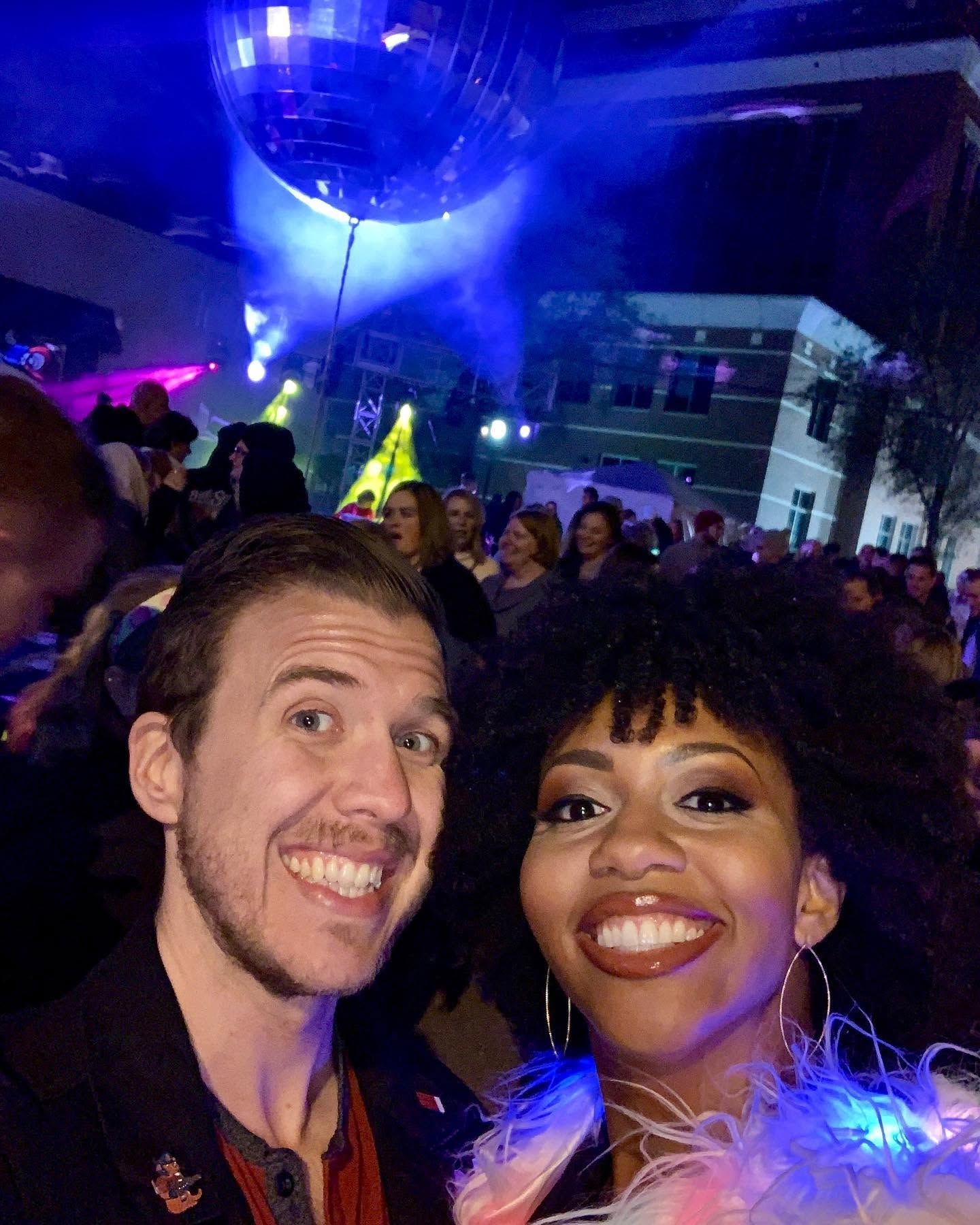 Two people smile for a selfie at a crowded night-out with a large disco ball above, purple and blue stage lights, and a building in the background.