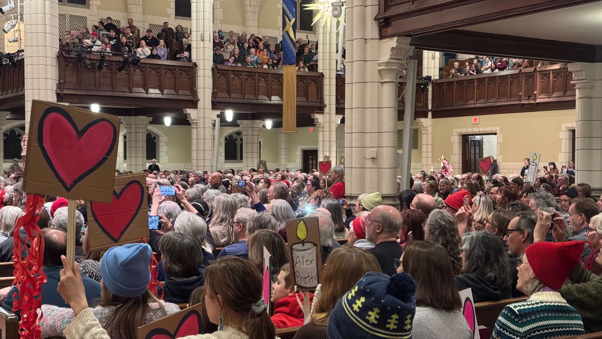 Large crowd inside a church holding handmade signs with red hearts and candles, attending a vigil or memorial service in a warmly lit, arched interior.