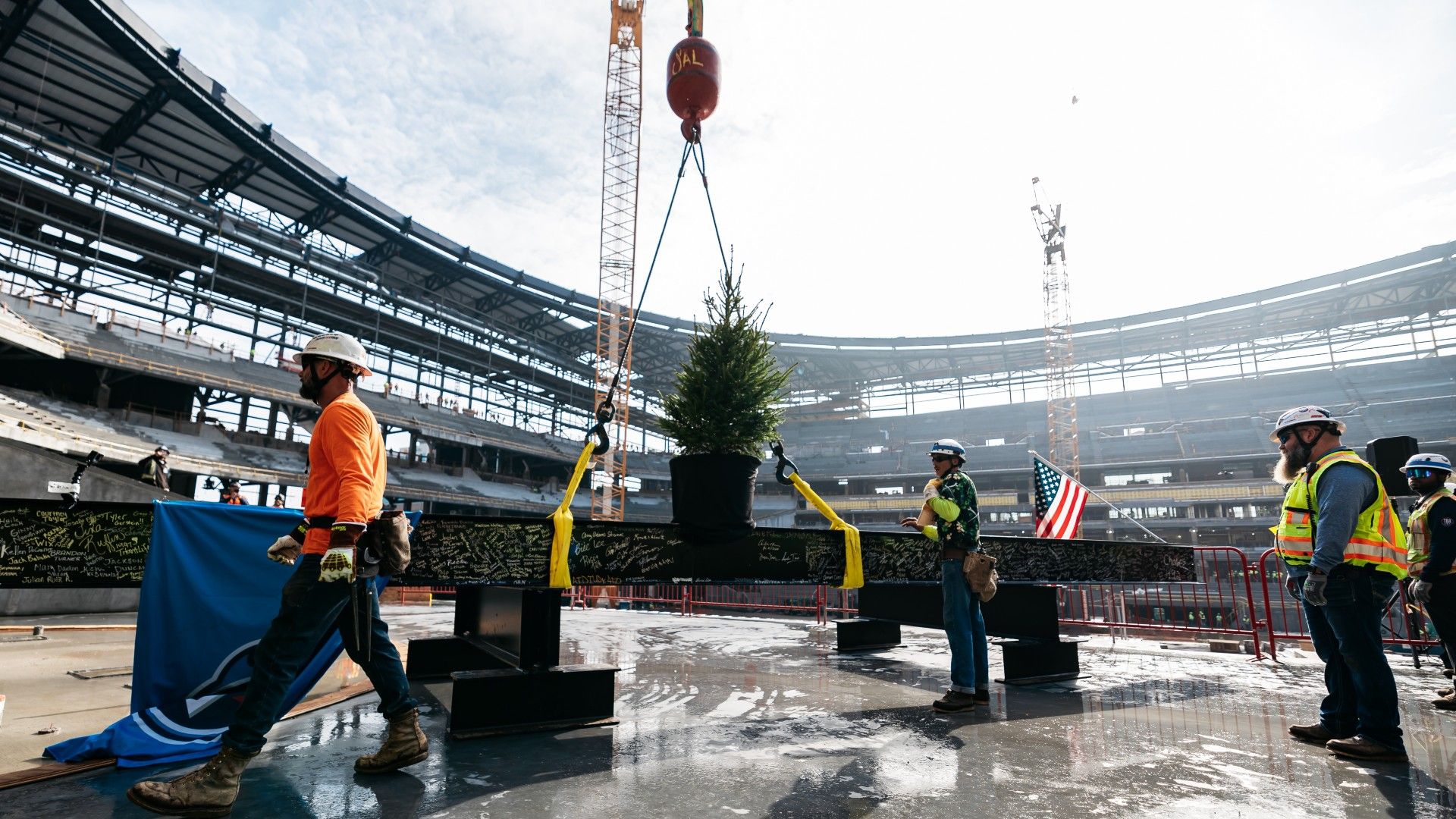 Construction workers wearing helmets and safety vests stand around a signed steel beam with a small Christmas tree attached, inside a large stadium under construction with cranes in background.