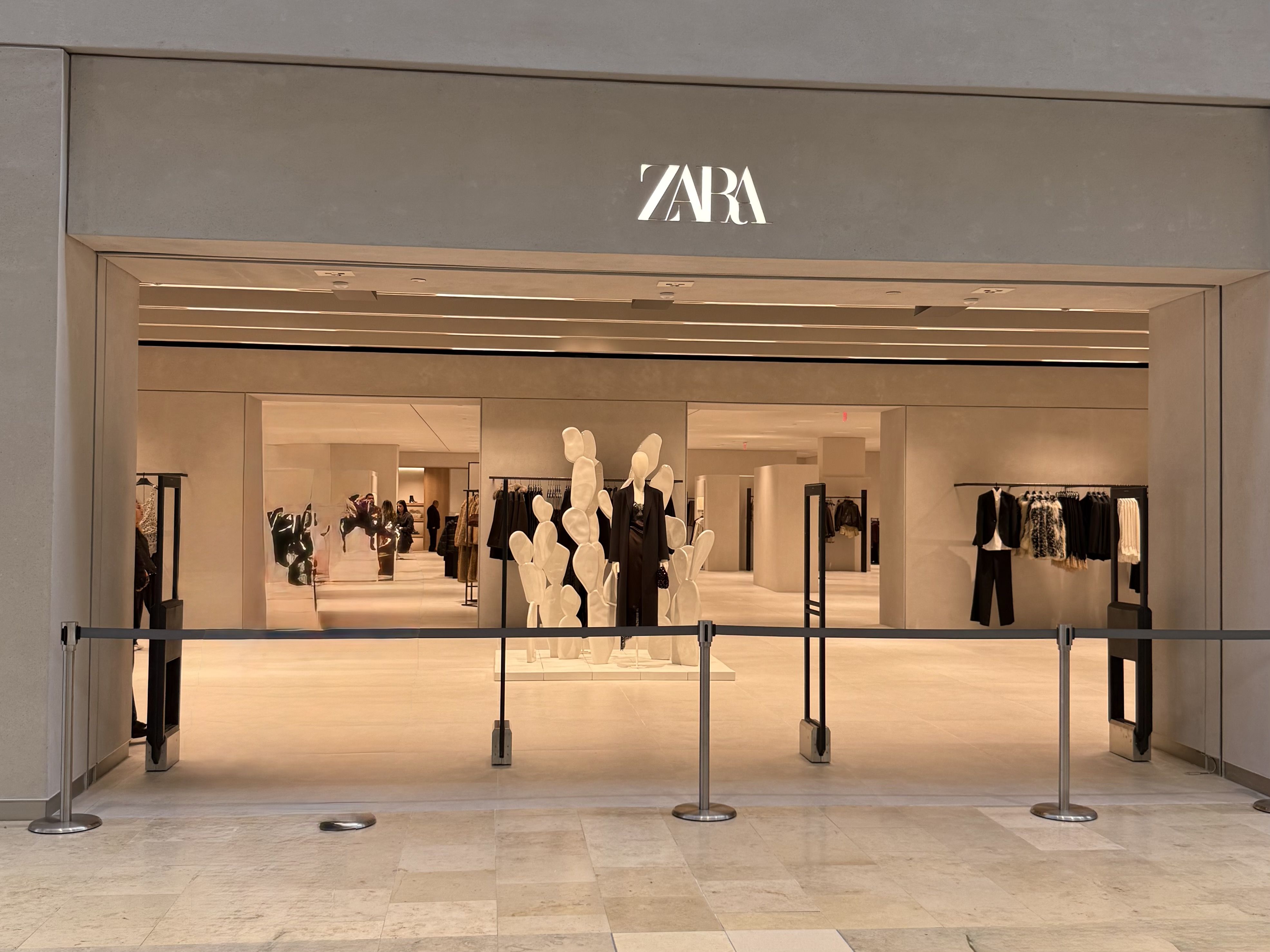 Entrance of a Zara store in a mall with a minimalist beige interior, mannequins dressed in black, and clothing racks; metal barriers block the entrance.